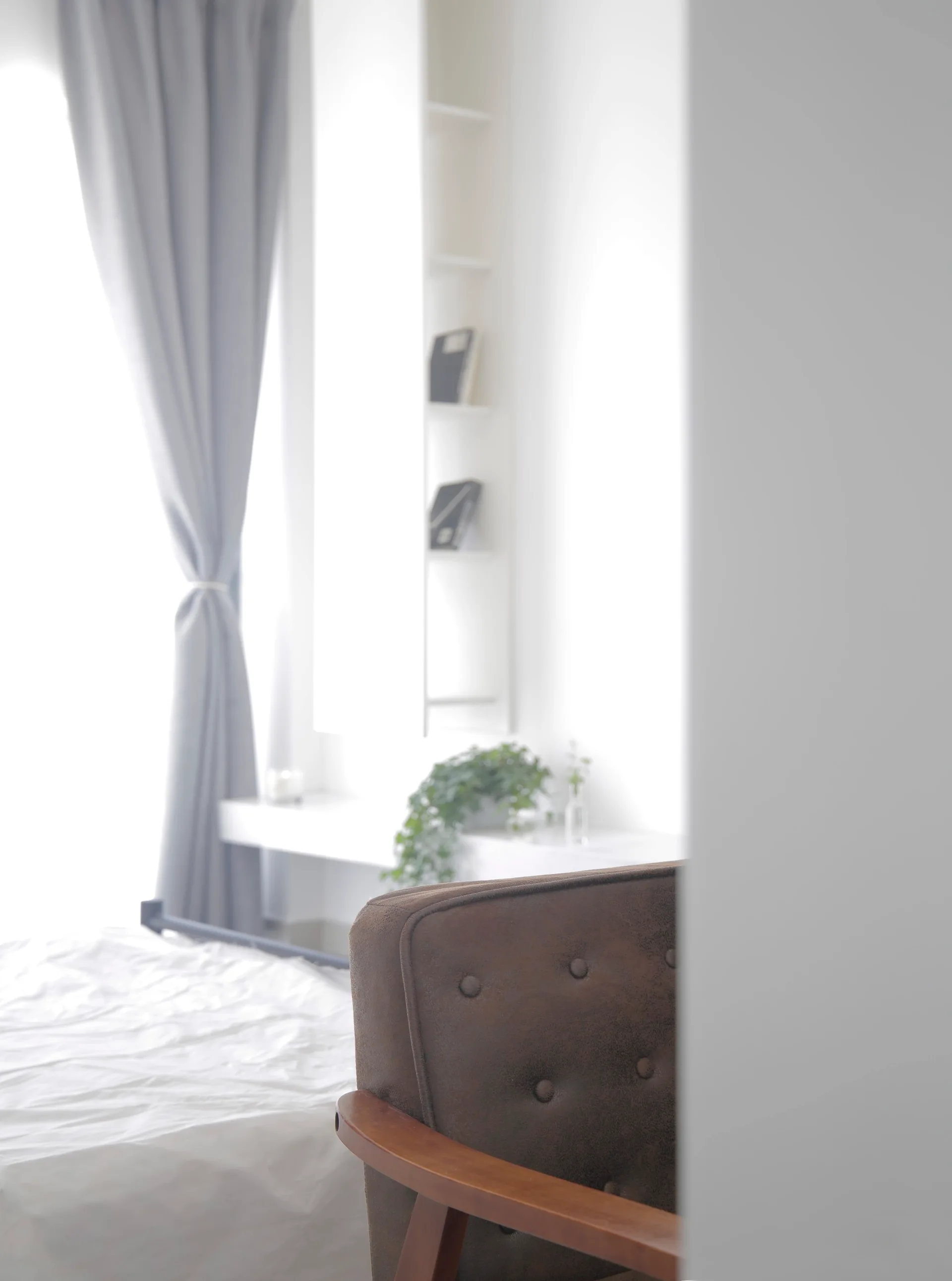 A partial view of a bedroom featuring a brown tufted armchair, white bedding, grey window curtains, and a floating white wall shelf.