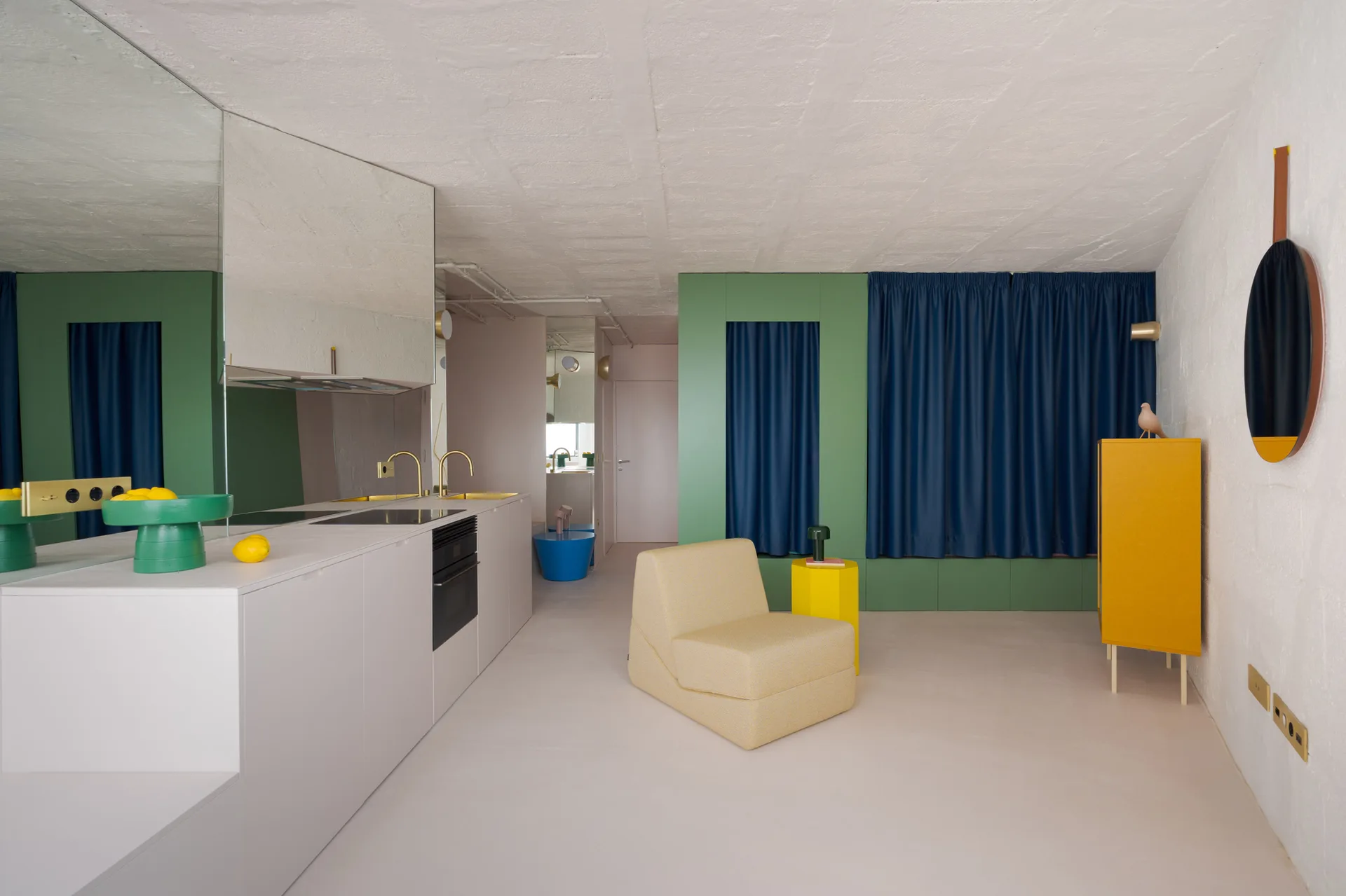 Minimalist white kitchen features flat-front cabinets, green wall paneling, deep navy curtains, and a yellow armchair on a seamless light floor.