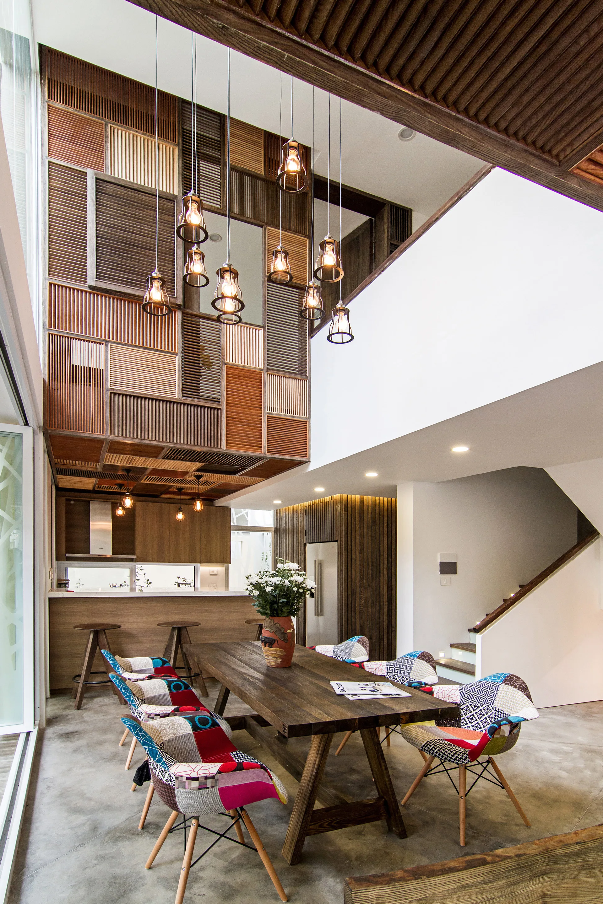 A double-height dining room features a long wooden table with colorful patchwork chairs under a cluster of hanging pendant lights.
