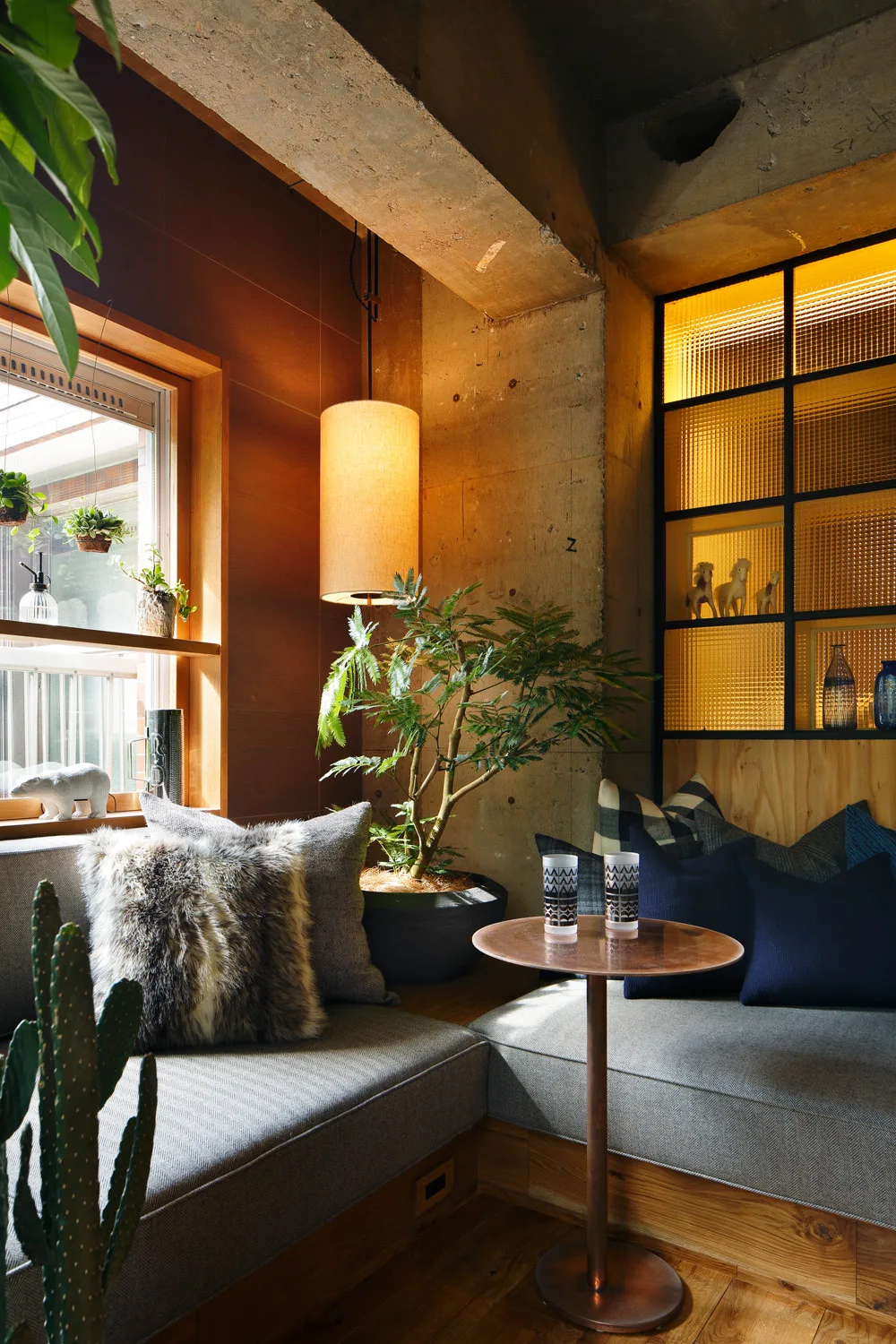 A cozy corner with L-shaped grey bench seating, a round copper pedestal table, an exposed concrete pillar, and a backlit textured glass partition.