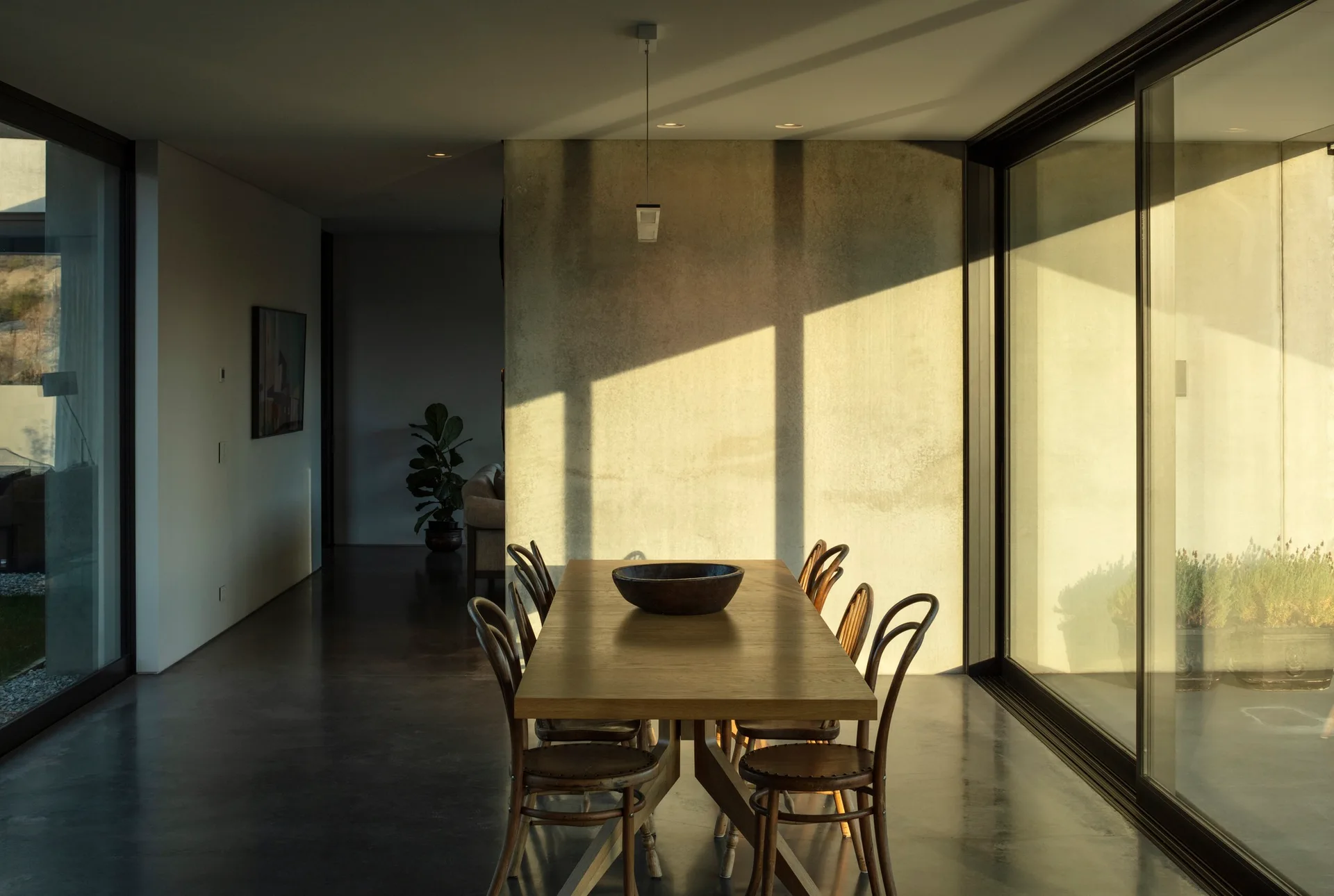 A long wooden dining table with curved wooden chairs stands next to large sliding glass doors and in front of a textured concrete wall.