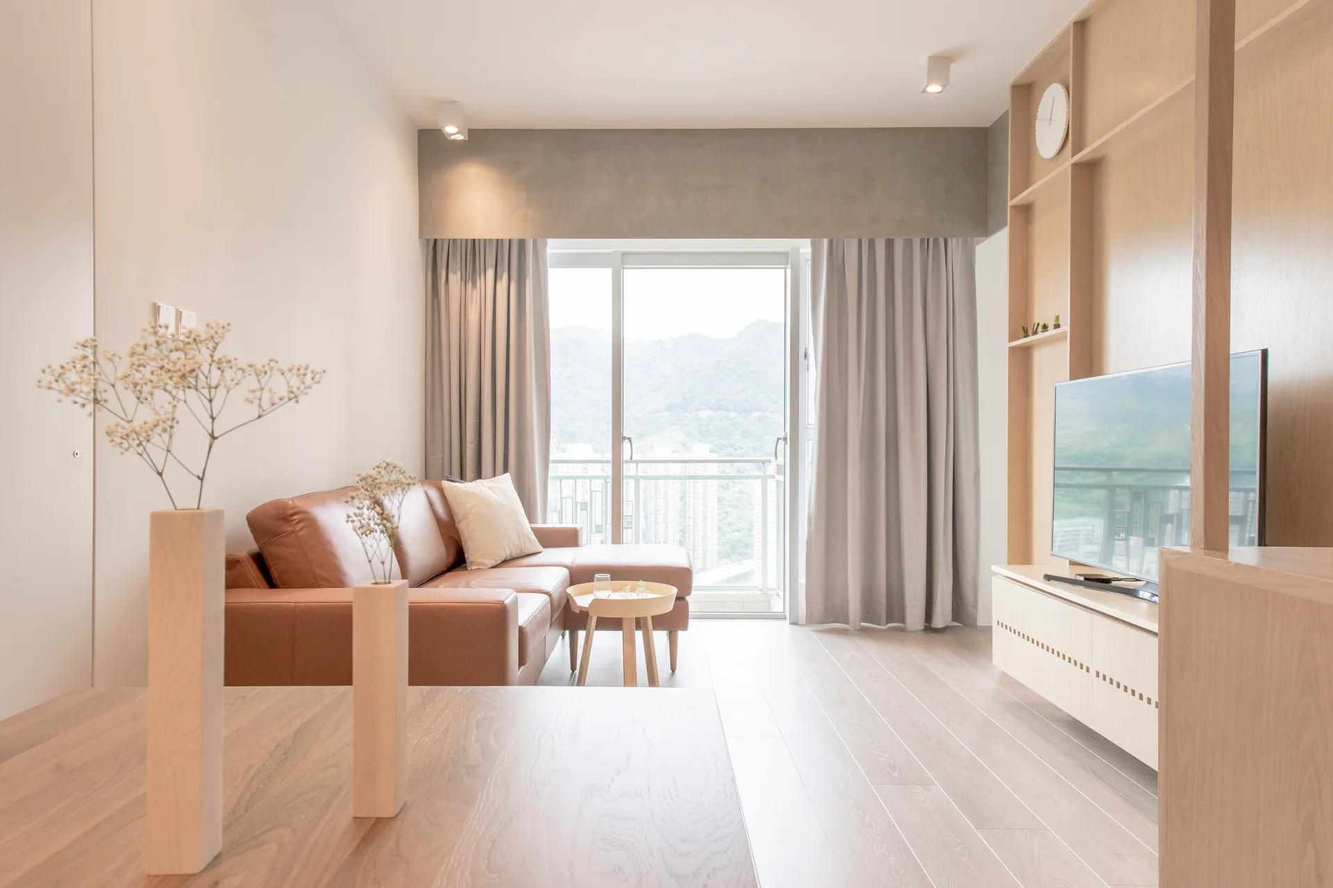 A minimalist living room features a brown leather sectional sofa, light wood media shelving, and grey curtains beside a large window.