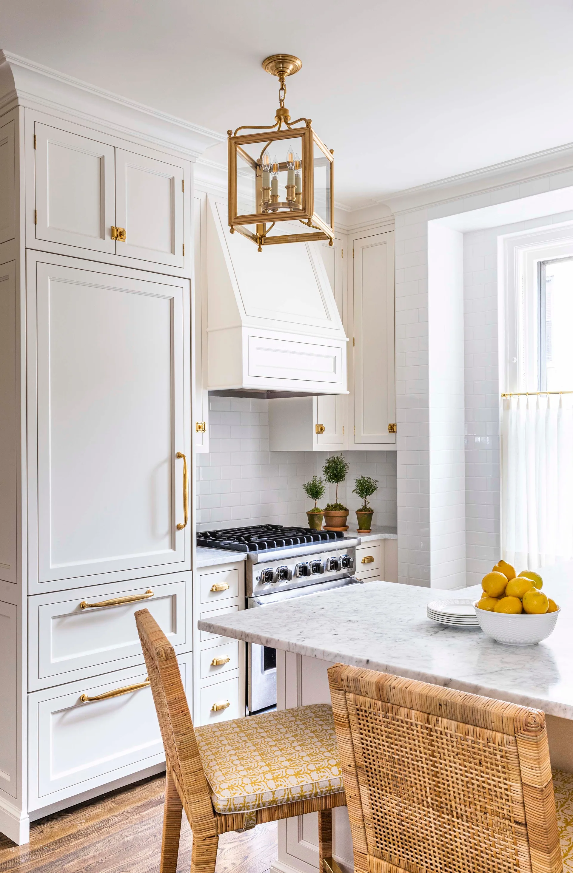 A white kitchen features paneled cabinetry with brass hardware, a marble-topped island, and two woven rattan stools with patterned cushions.