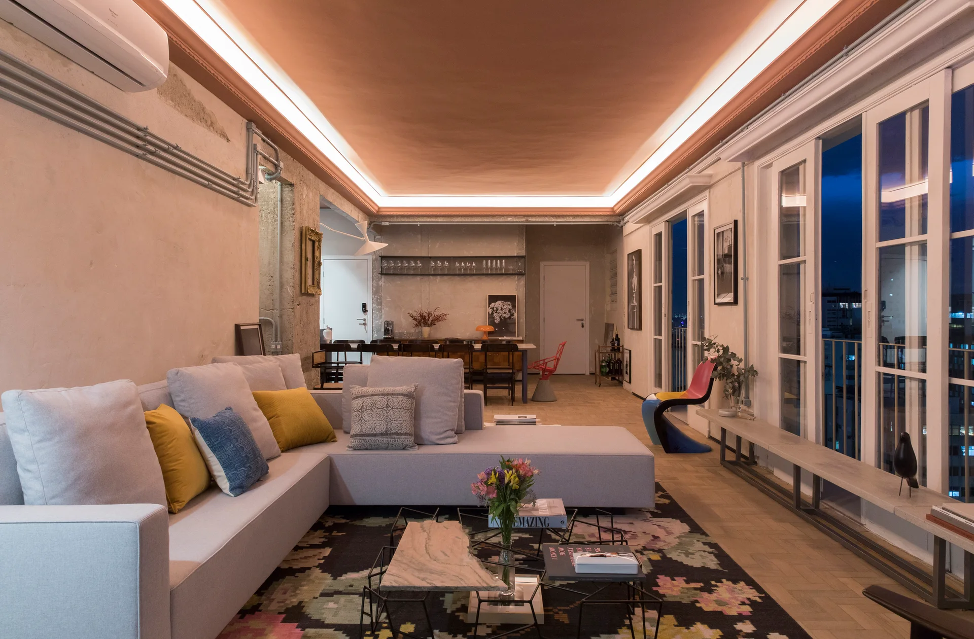 An industrial living room features a grey sectional sofa on a patterned rug beneath a warm terracotta ceiling with integrated cove lighting.