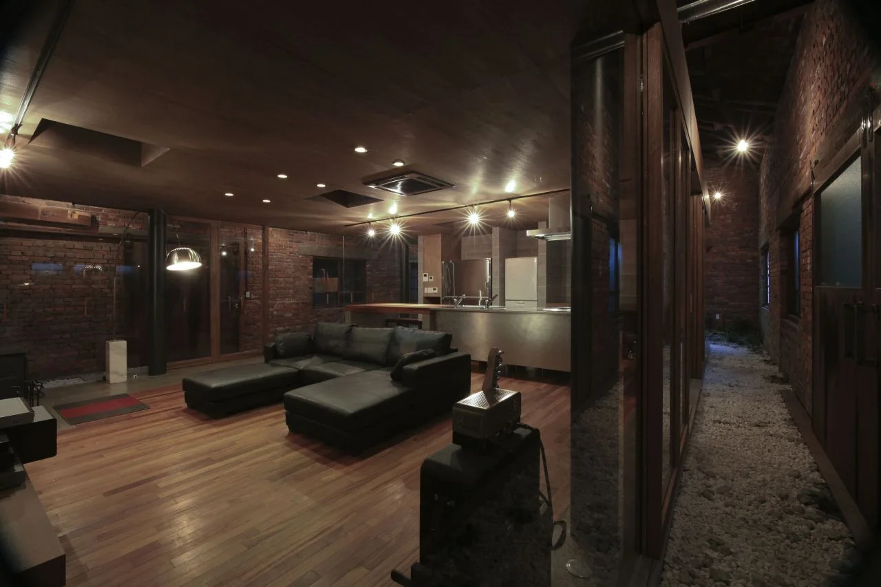 An industrial living room features a black leather sectional sofa, exposed brick walls, warm wood flooring, and a glass-enclosed indoor gravel path.