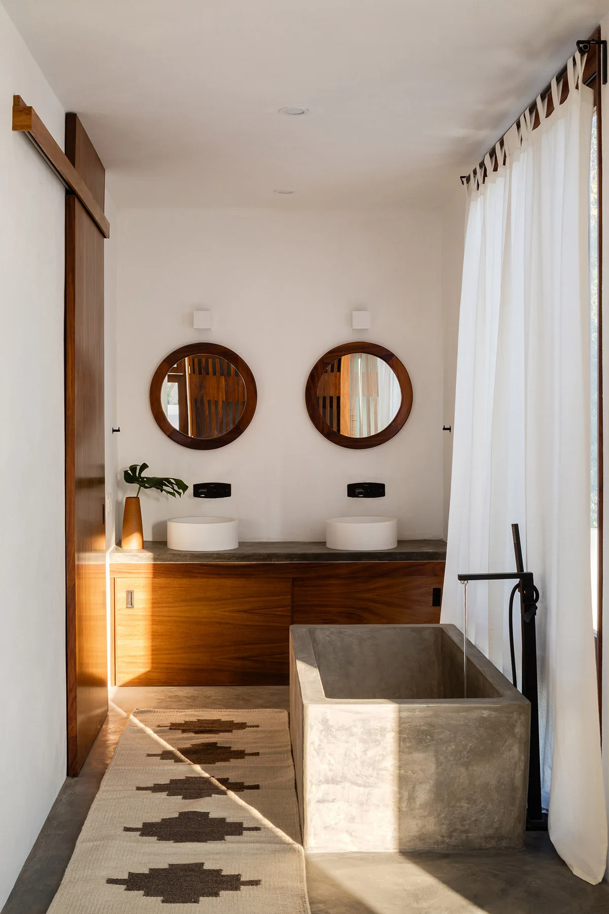 An organic modern bathroom featuring a rectangular concrete soaking tub, a wooden double vanity with round mirrors, and a patterned runner rug.