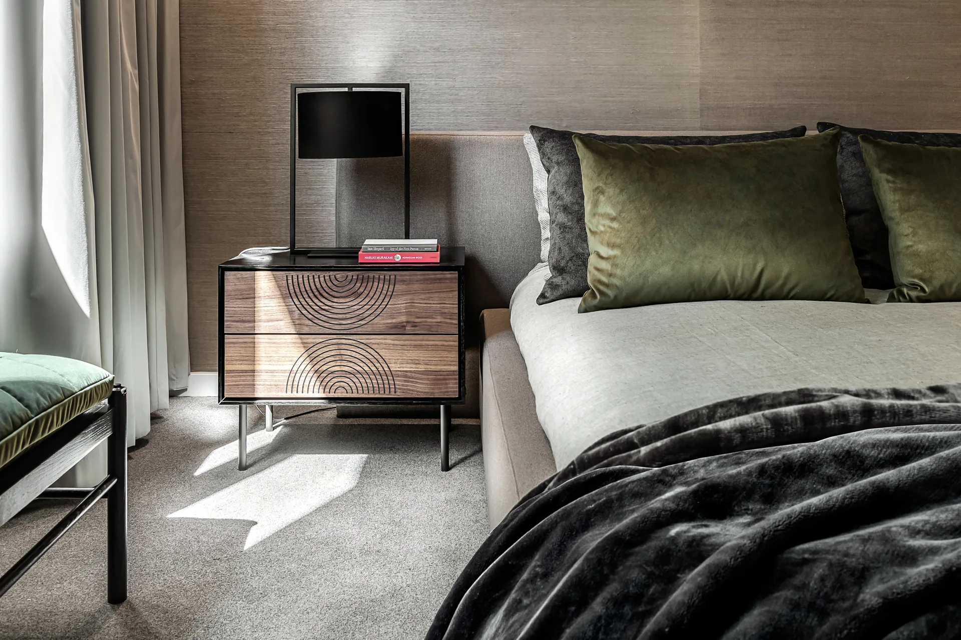 A contemporary bedroom featuring an upholstered bed with green and grey cushions next to a carved wood nightstand with a black lamp.