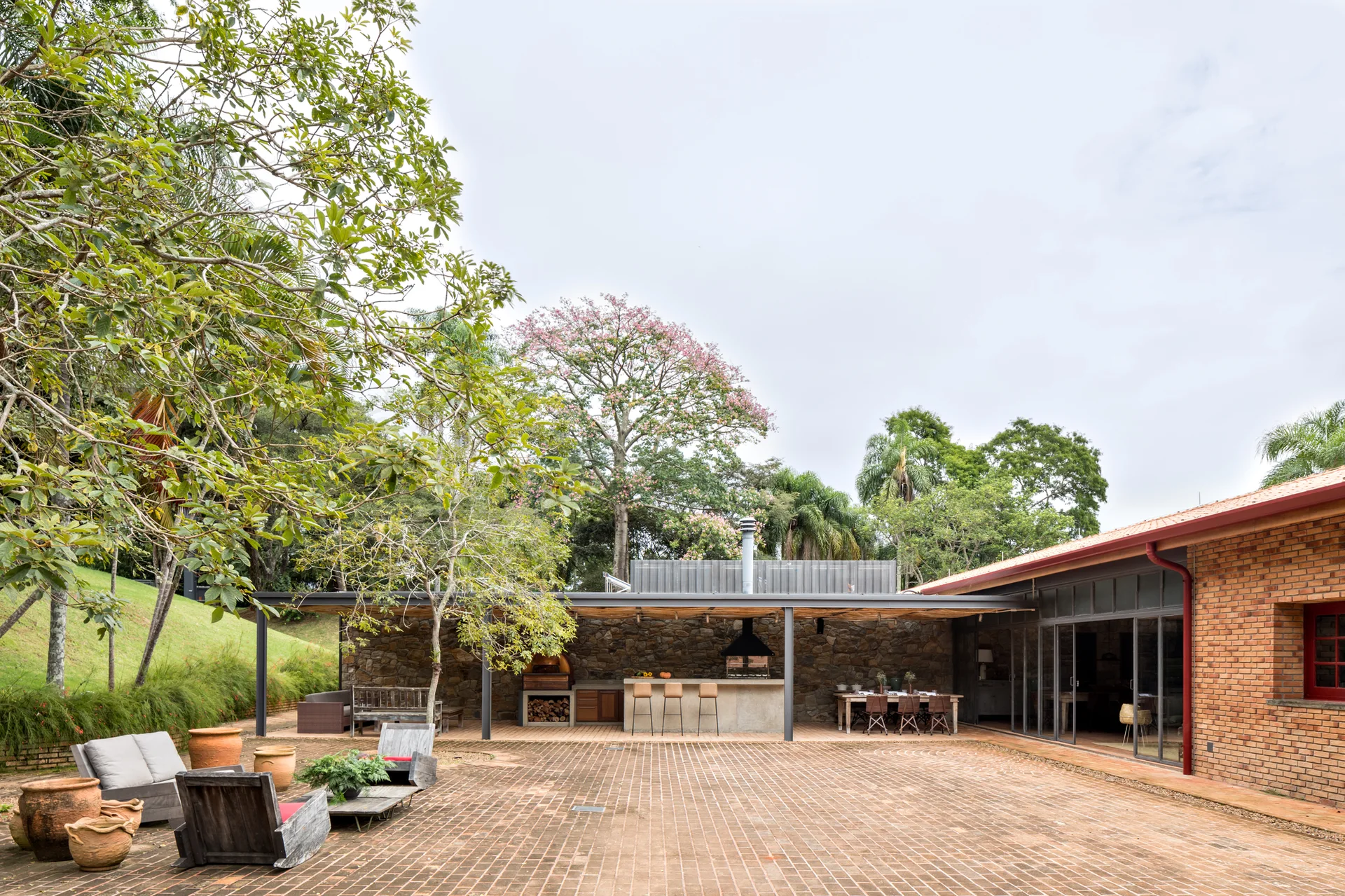 A spacious brick-paved patio features a covered outdoor kitchen with a stone back wall, a dining table, and casual lounge chairs with terracotta planters.