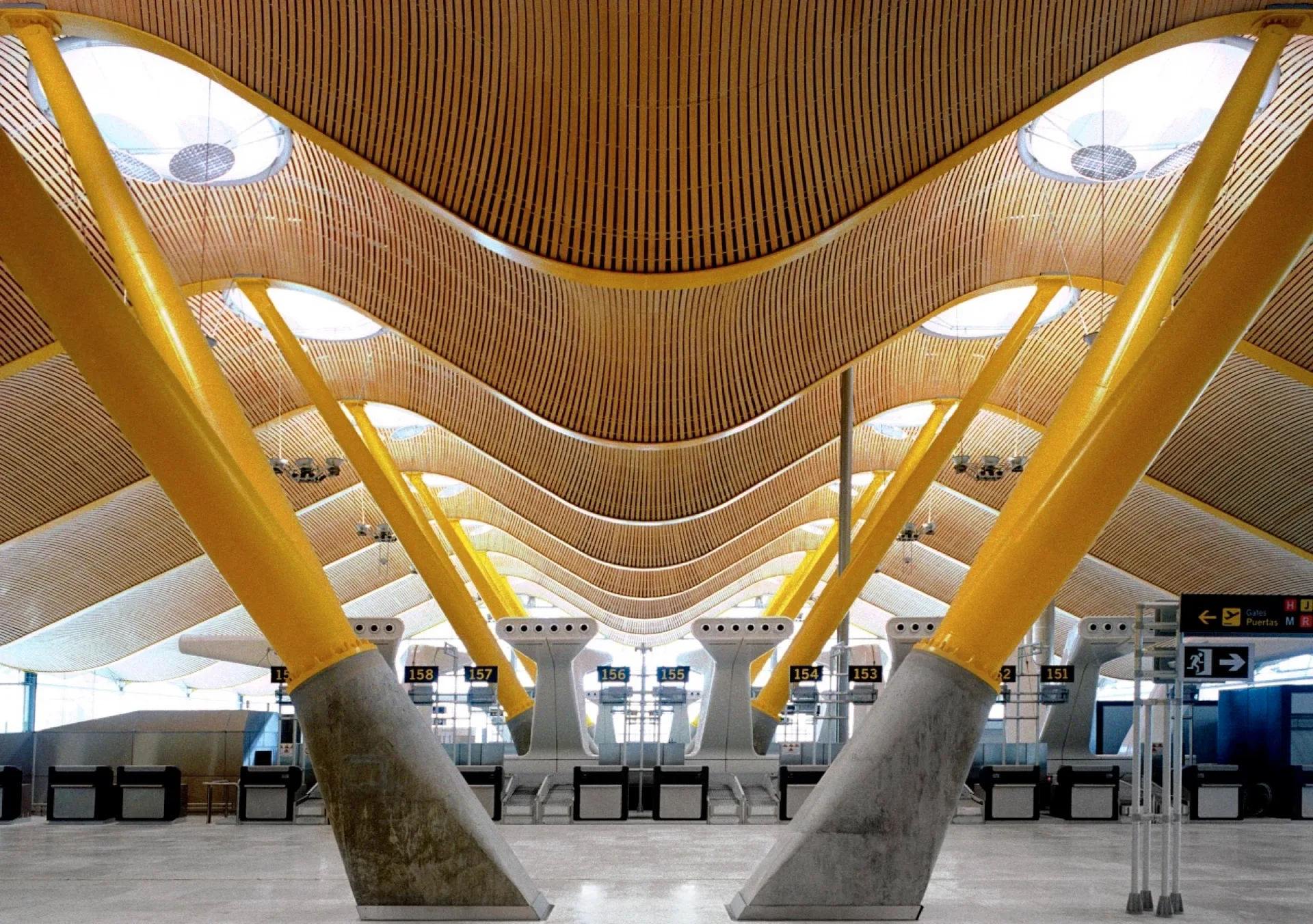 A spacious terminal interior features an undulating slatted wood ceiling supported by angled yellow steel columns with heavy concrete bases.