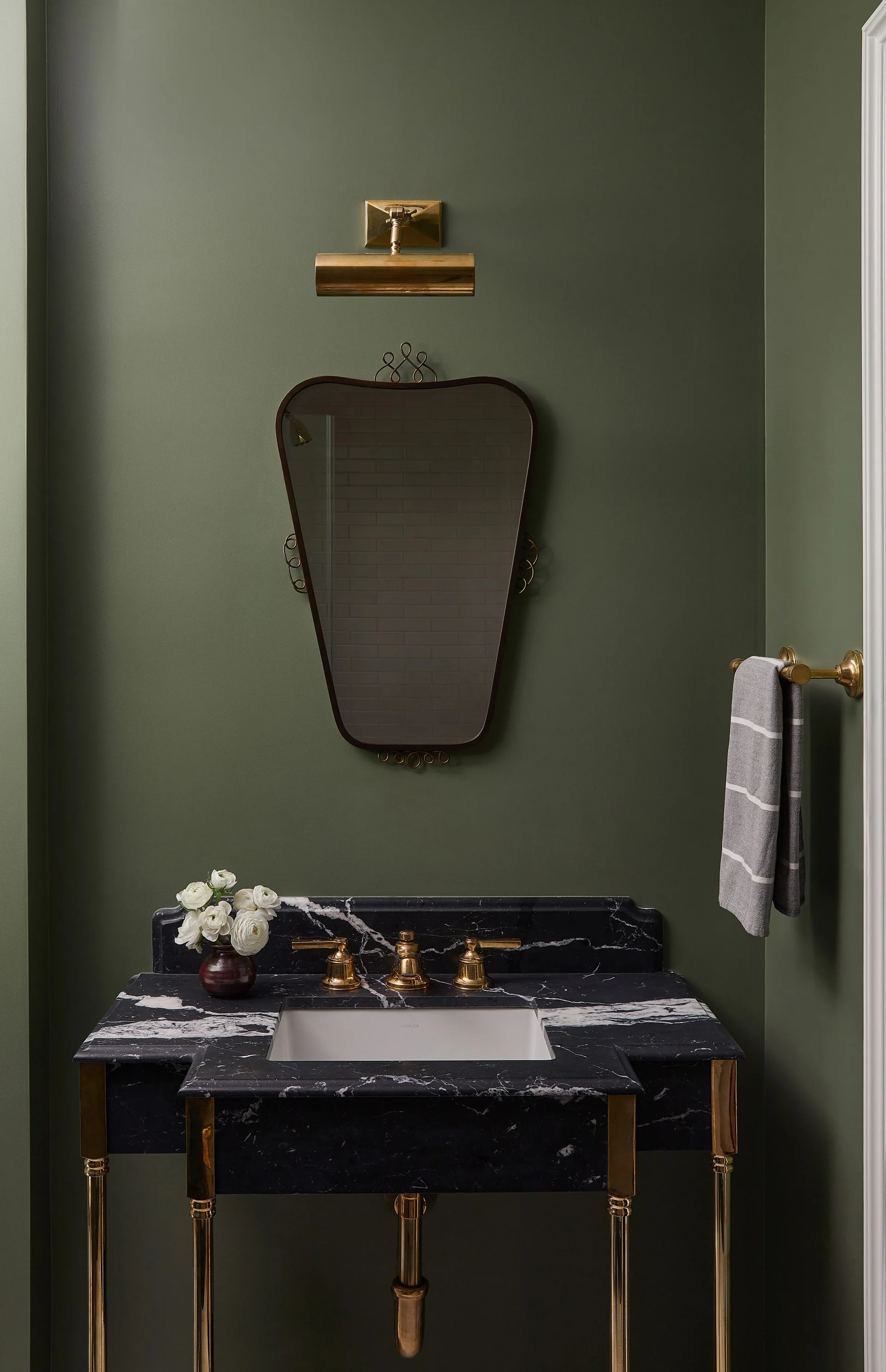 An olive green powder room features a black marble console sink, a trapezoidal wall mirror, and brass hardware including a picture light and faucet.