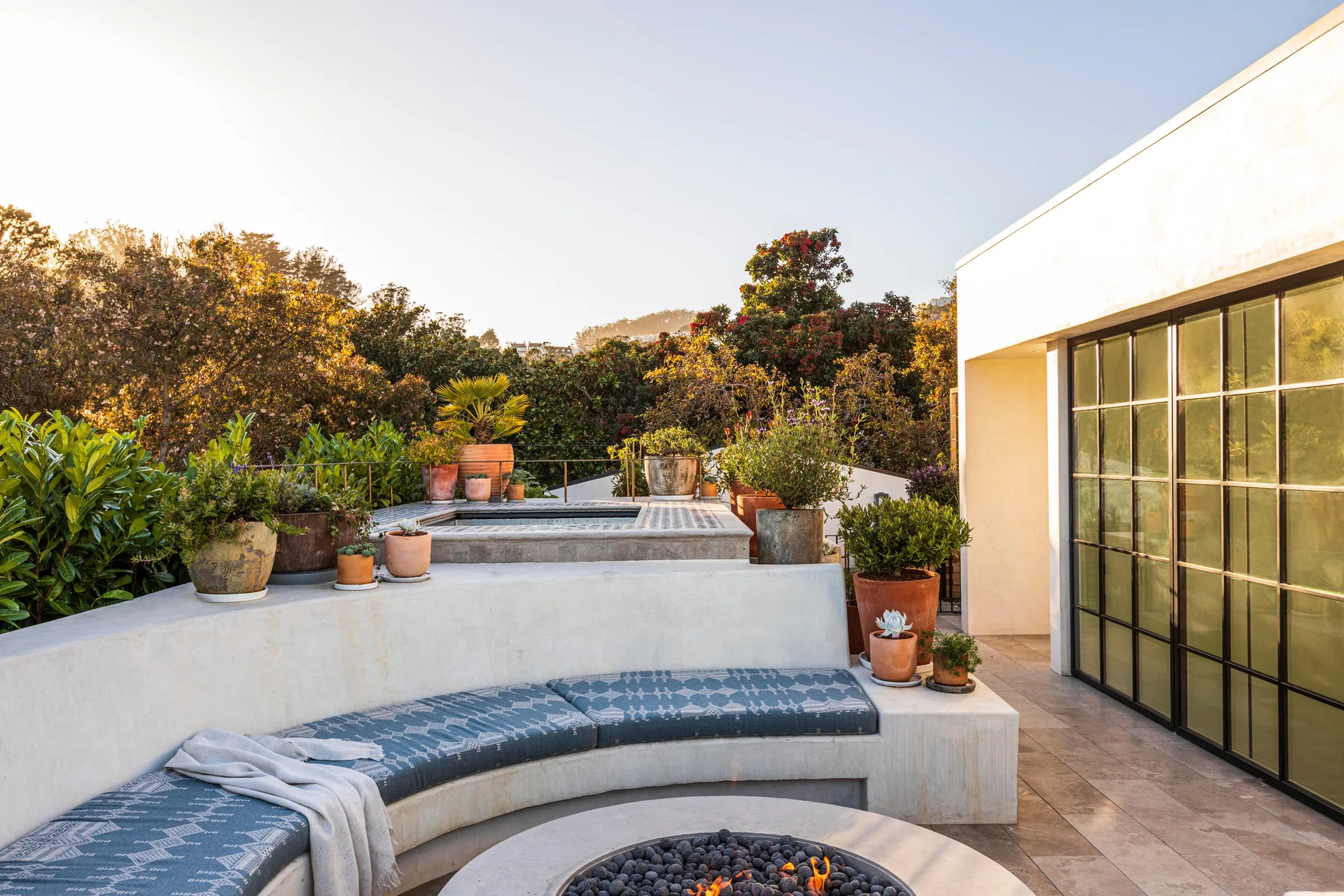 An outdoor concrete patio featuring a curved built-in bench with blue patterned cushions, a fire pit, and various potted plants.