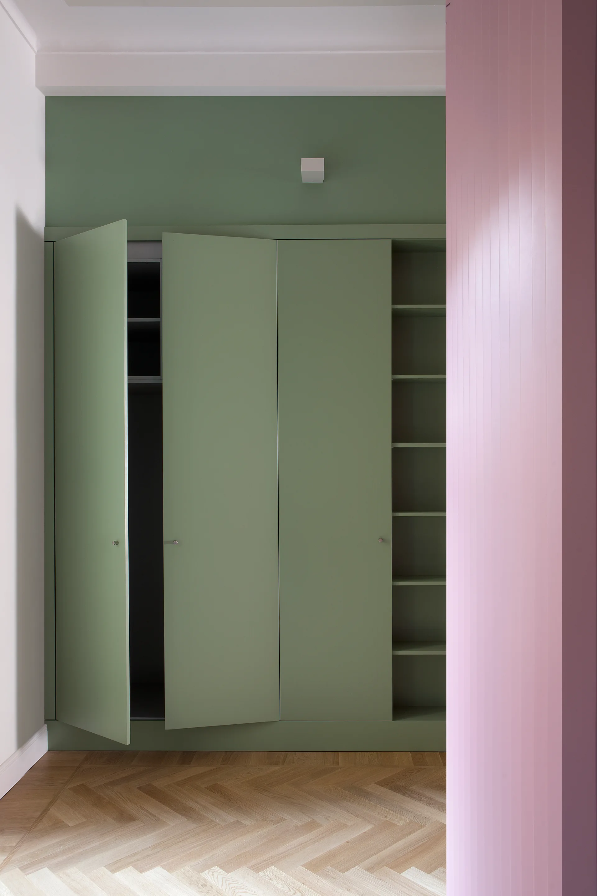 A built-in sage green wardrobe with open and closed shelves stands on herringbone wood flooring next to a pink slatted wall panel.
