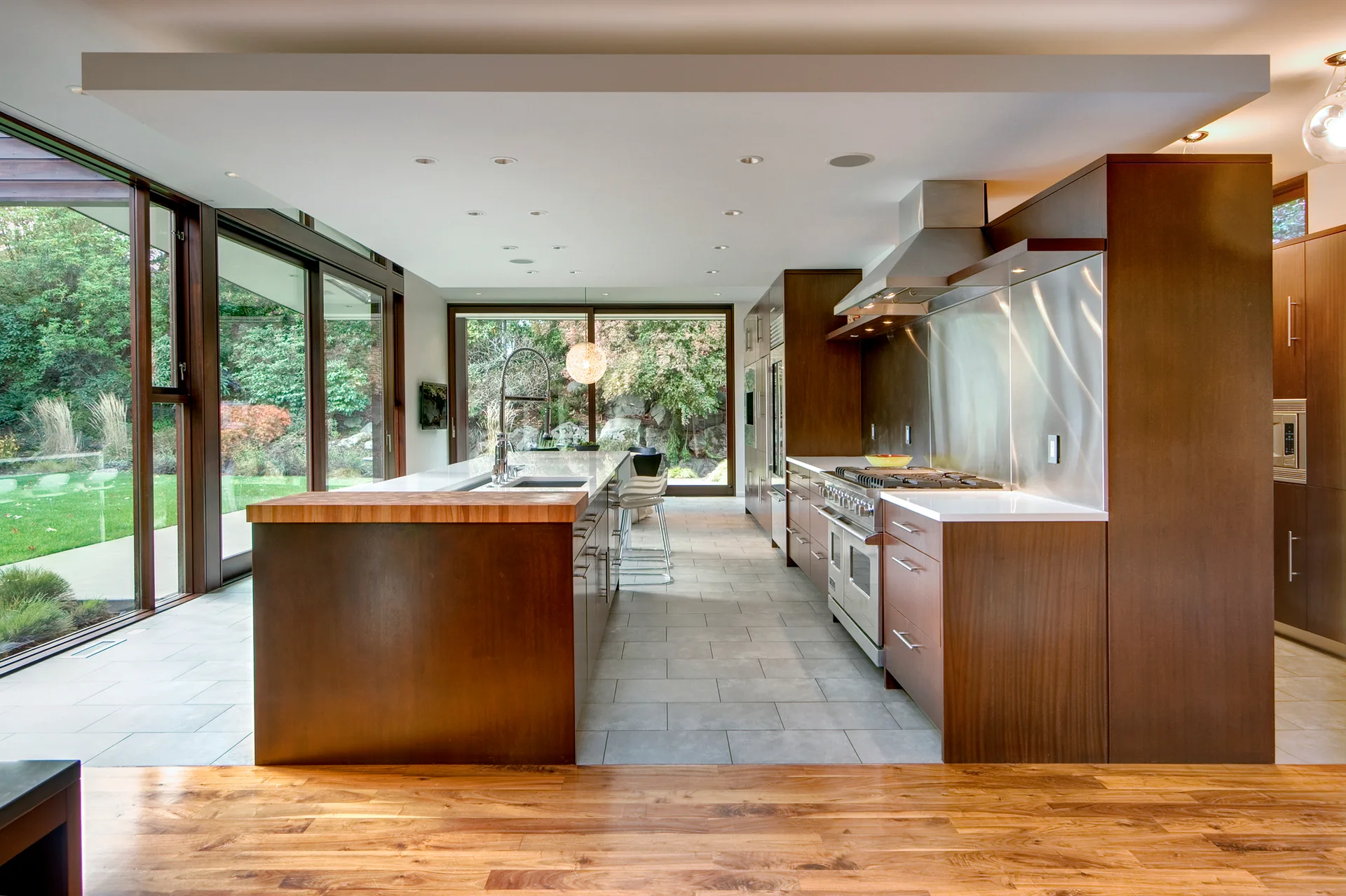 A contemporary kitchen with dark wood cabinets, a large central island, and floor-to-ceiling windows showing a lush green garden.