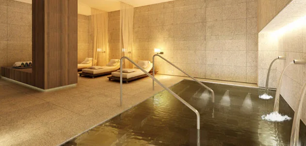 An indoor spa pool featuring metal railings, wall-mounted water spouts, stone tile walls, and modern lounge chairs with sheer curtains.