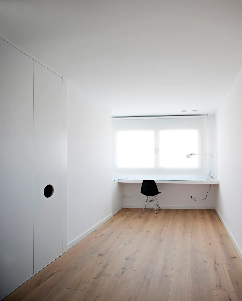 A minimalist white workspace with a wall-mounted desk, black task chair, white built-in closets, and light oak hardwood floors.