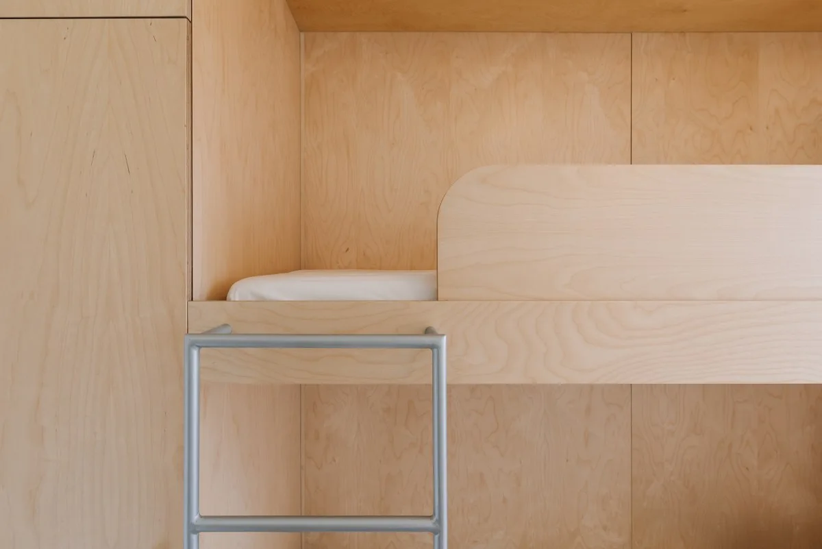 A minimalist built-in bunk bed crafted from light plywood features a curved guardrail, a white mattress, and a grey metal ladder.