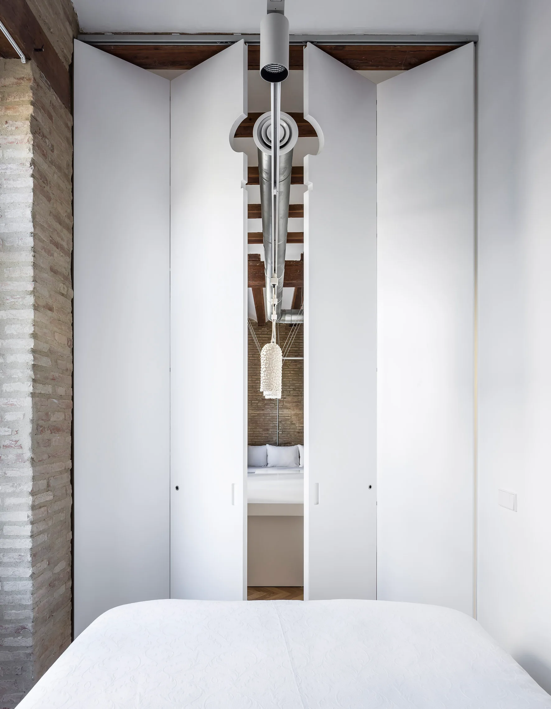 Large white bi-fold doors with a custom top cutout for industrial ductwork divide a bedroom with exposed brick walls and ceiling beams.