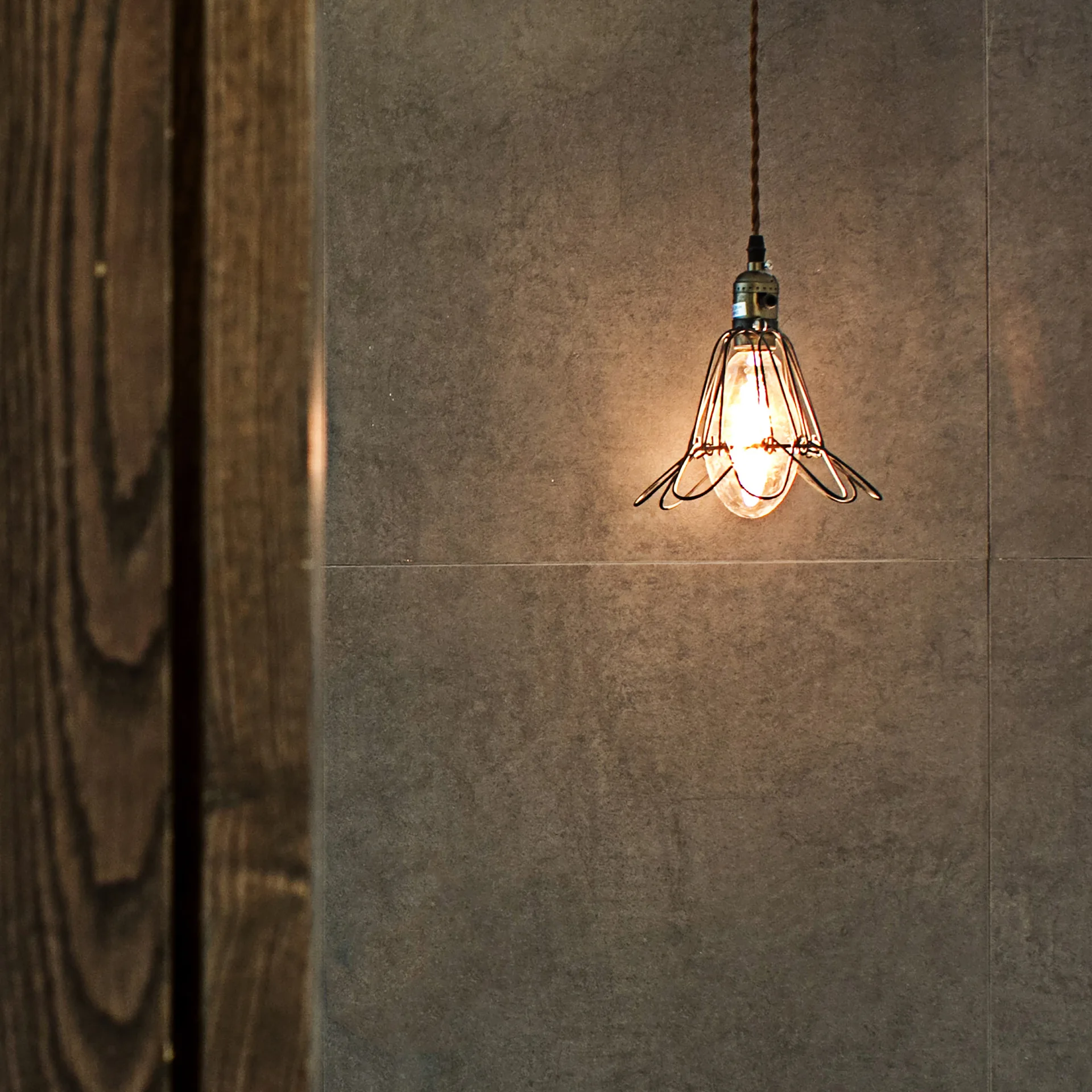 An industrial wire cage pendant light with a warm glowing bulb hangs against a matte grey tiled wall next to a dark wood panel.