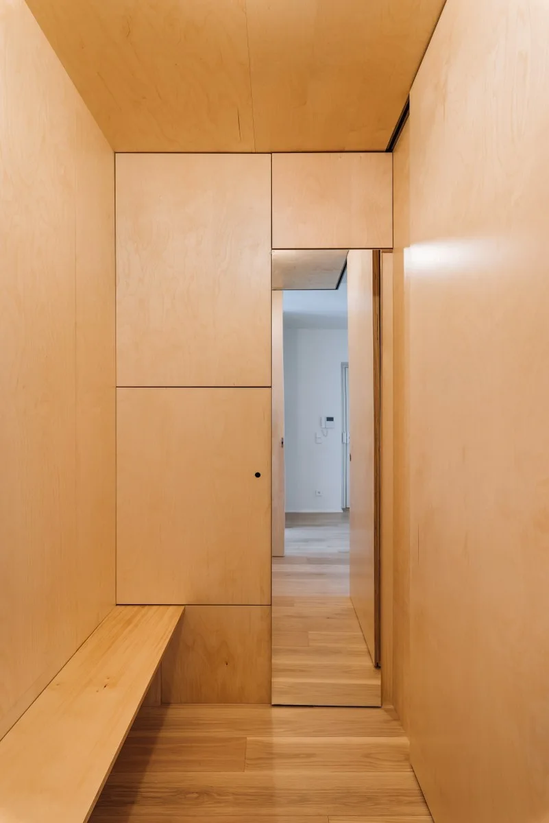 A narrow entryway featuring floor-to-ceiling light plywood paneling, an integrated wood bench, and seamless built-in closet doors.