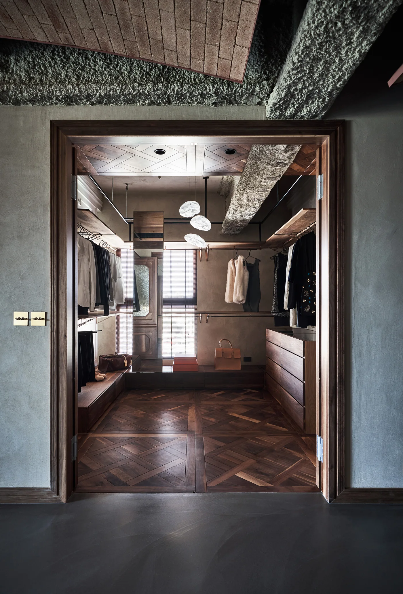 A walk-in closet framed by a wooden doorway features geometric parquet flooring, custom dark walnut cabinetry, and an exposed concrete ceiling beam.