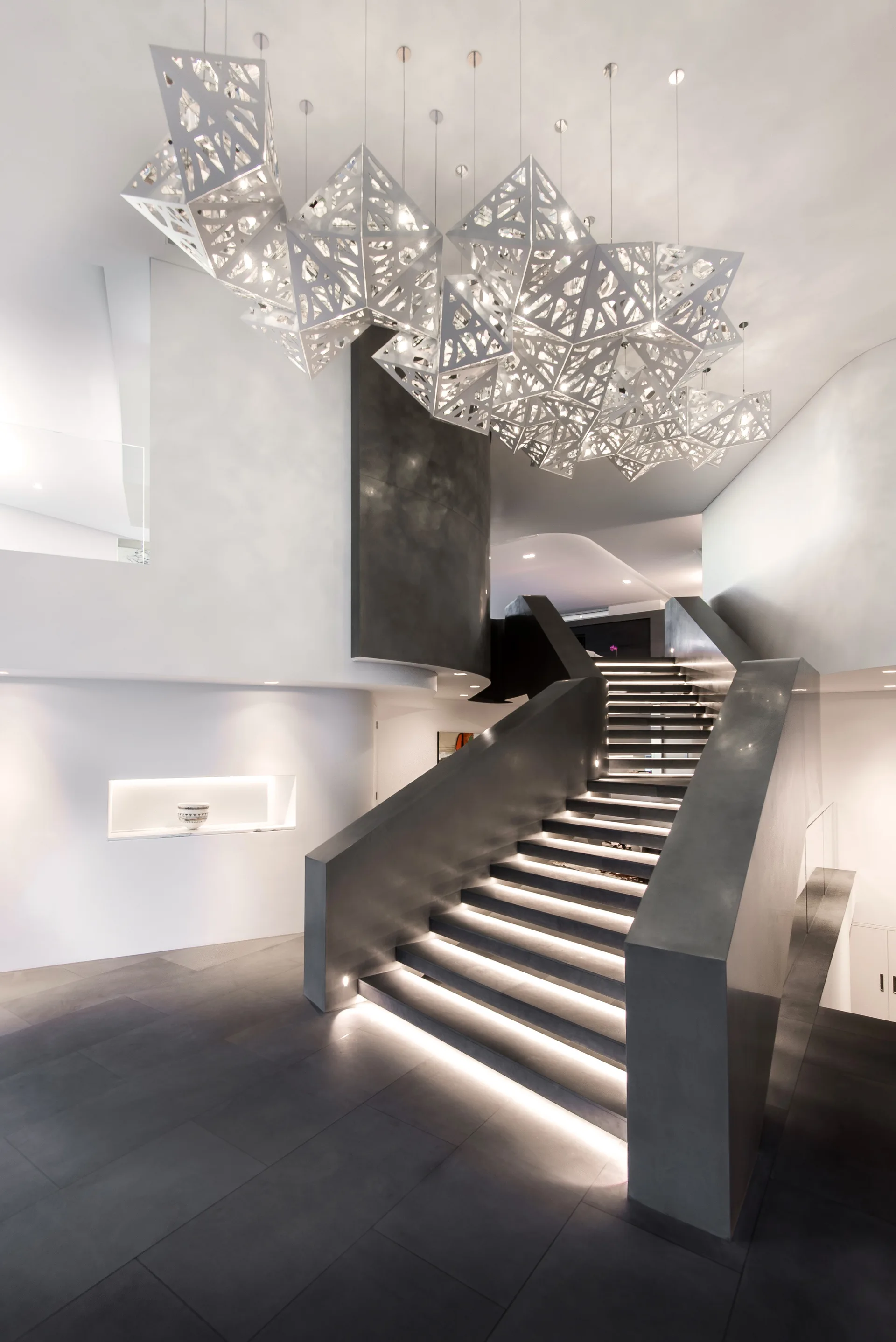 A dark modern staircase with integrated tread lighting sits beneath a large, faceted geometric pendant light fixture in a white-walled foyer.