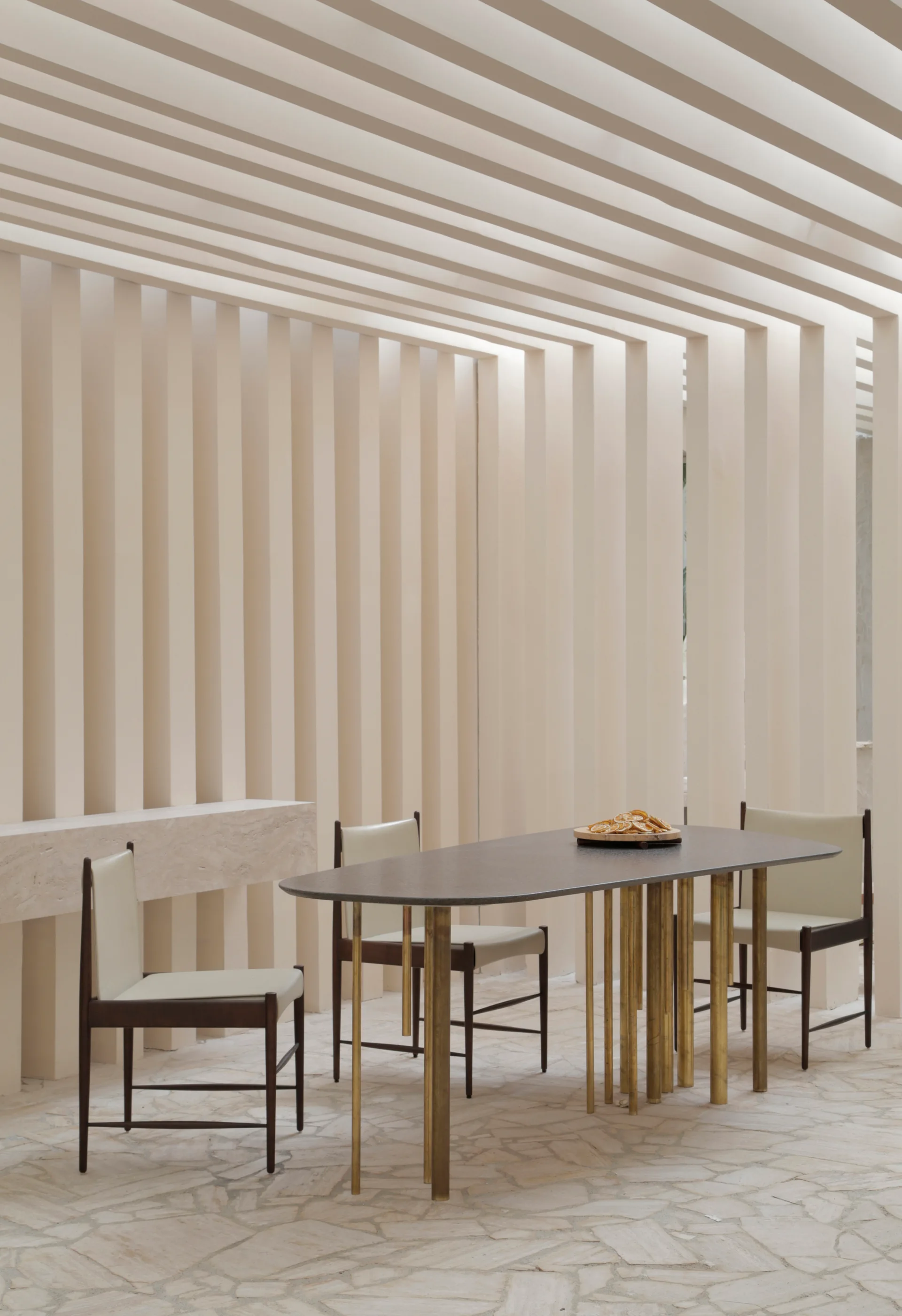 An oval dining table with gold legs and leather chairs stands on a flagstone floor beneath a continuous slatted cream wall and ceiling structure.