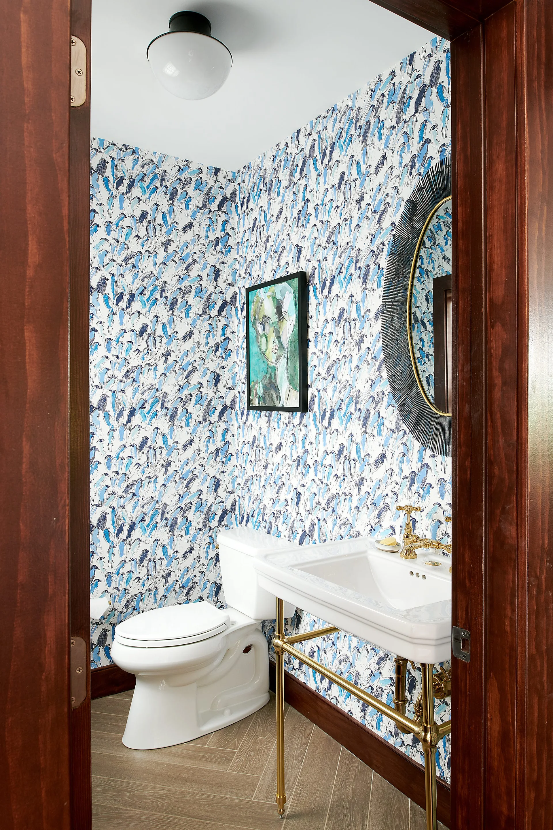 A powder room viewed through a dark wood doorway featuring blue and white patterned wallpaper, a white toilet, and a sink with a brass stand.