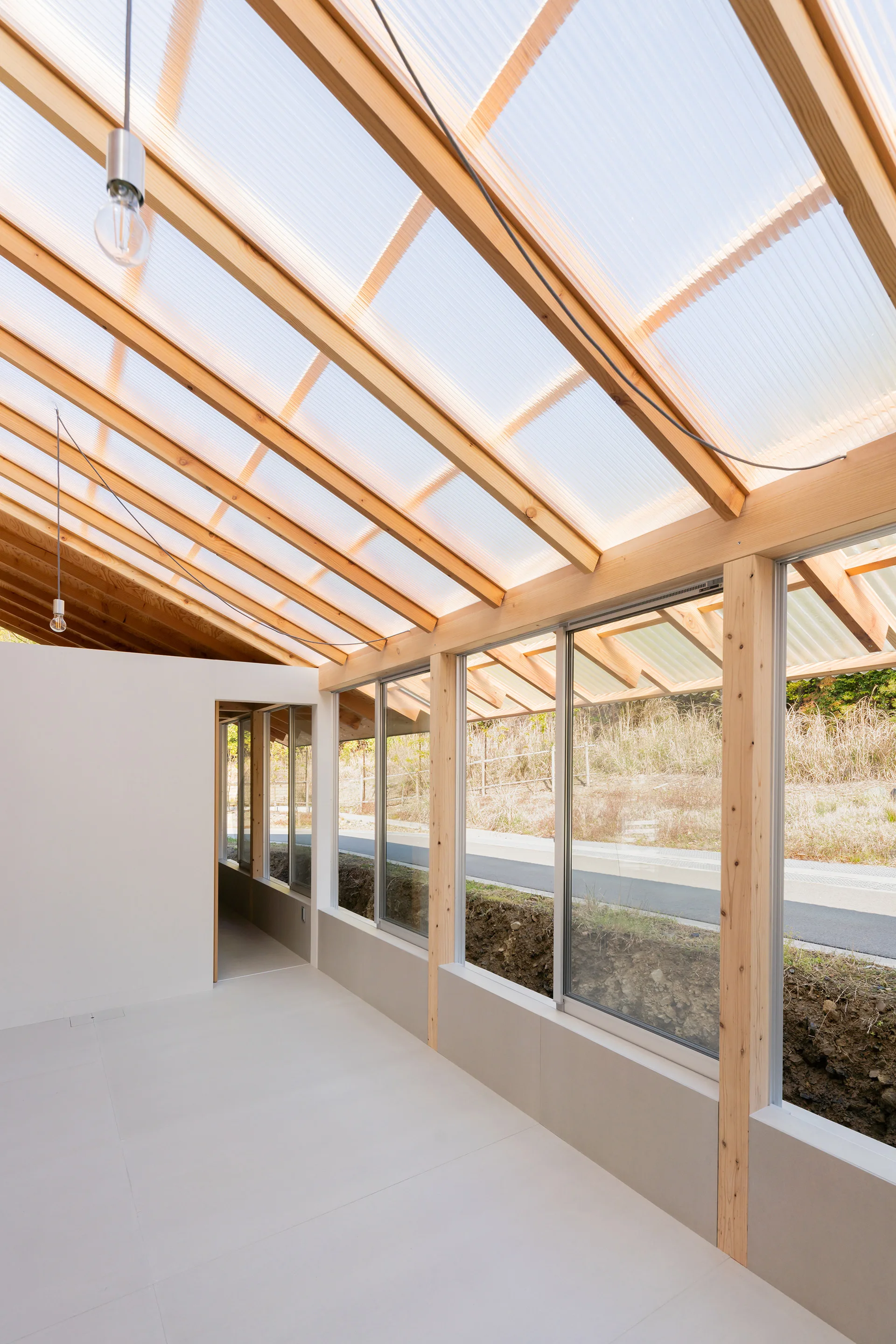 A minimalist hallway features an exposed light wood timber frame, large glass windows, and a sloped translucent polycarbonate roof.