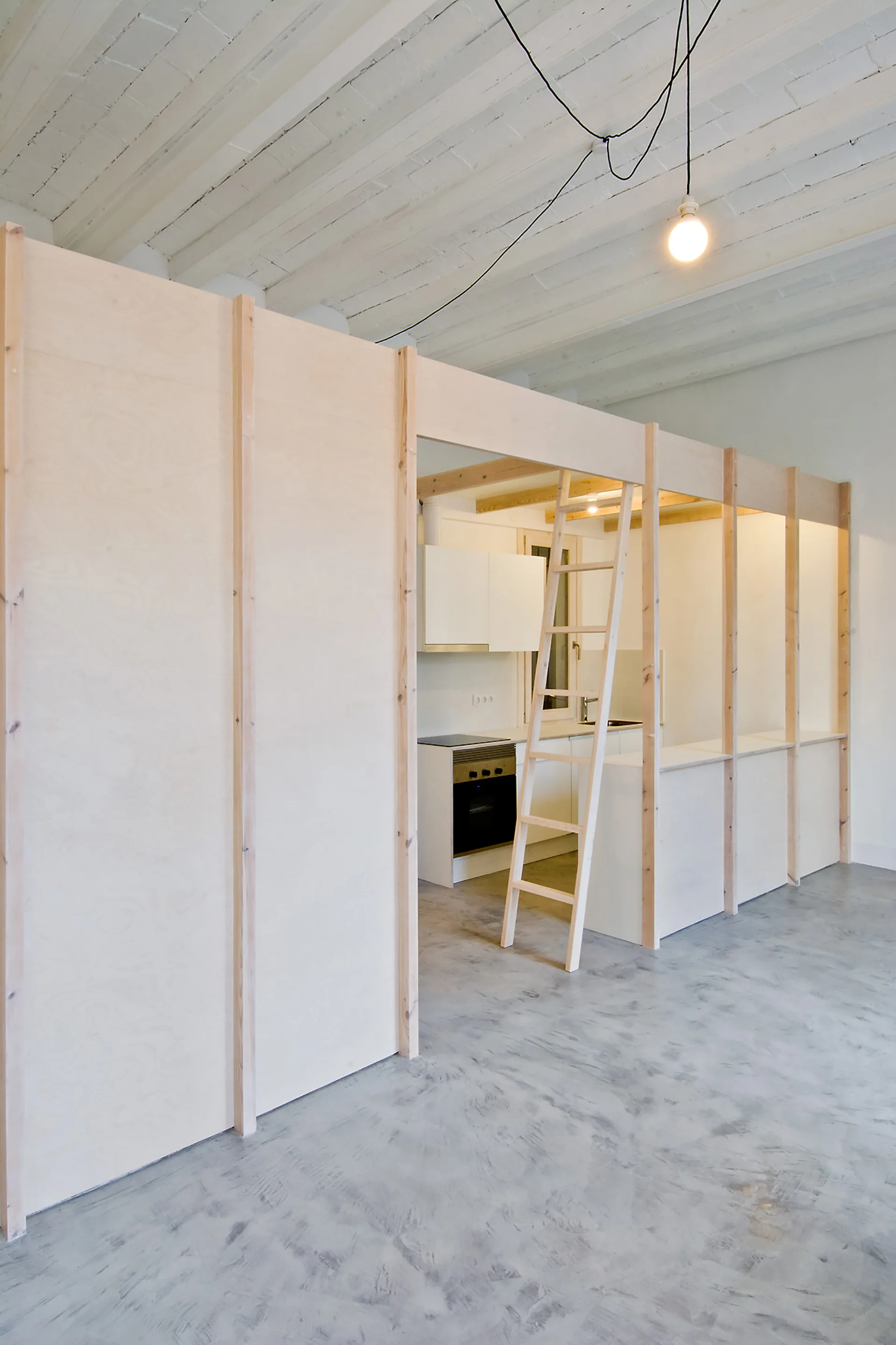 A light-colored plywood partition wall with a leaning wooden ladder stands on a grey concrete floor, separating a kitchen with white cabinets.