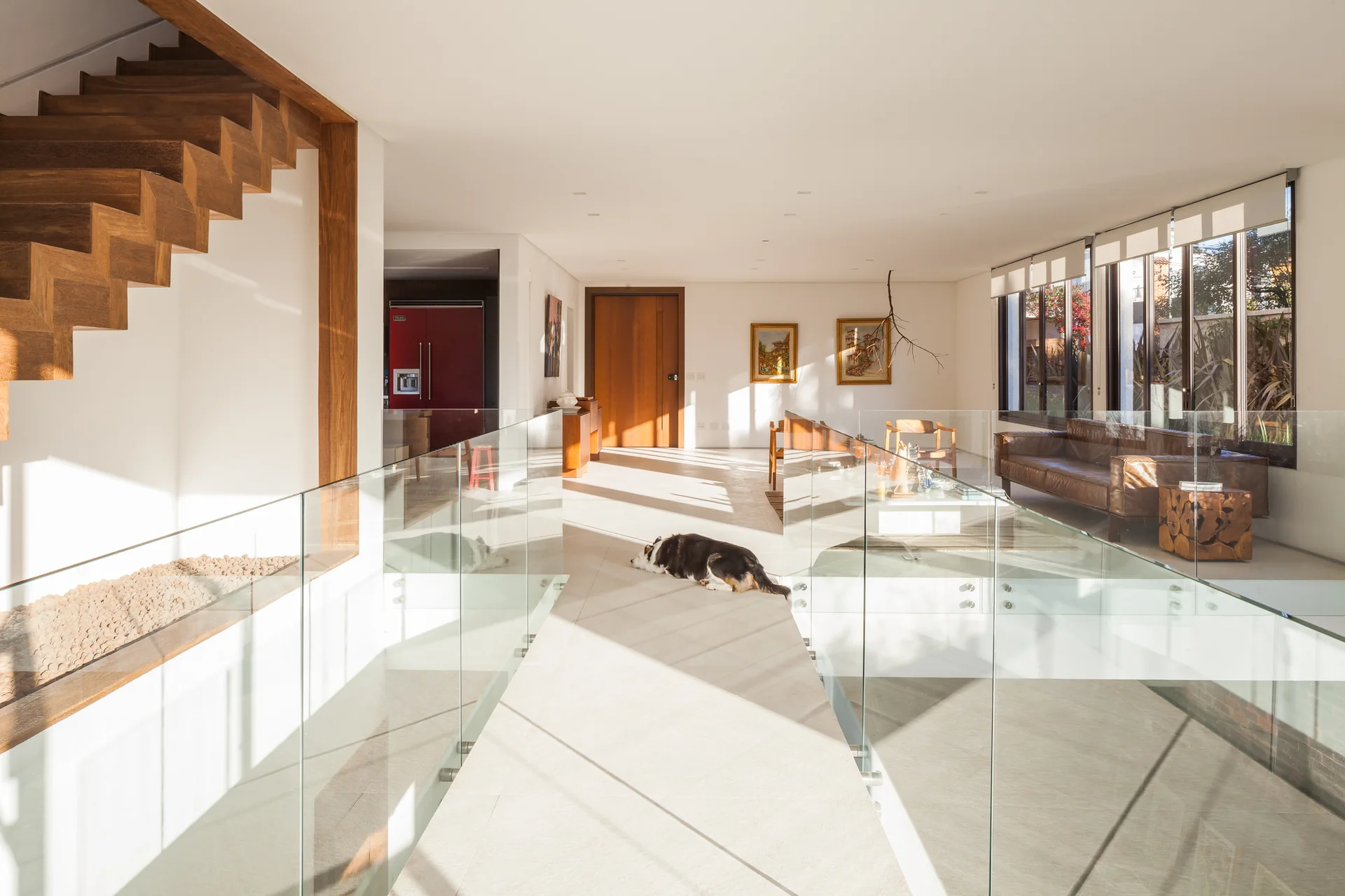 A bright upper-level landing featuring a wood staircase, transparent glass railings, beige tiled floors, and a sunlit living room in the background.