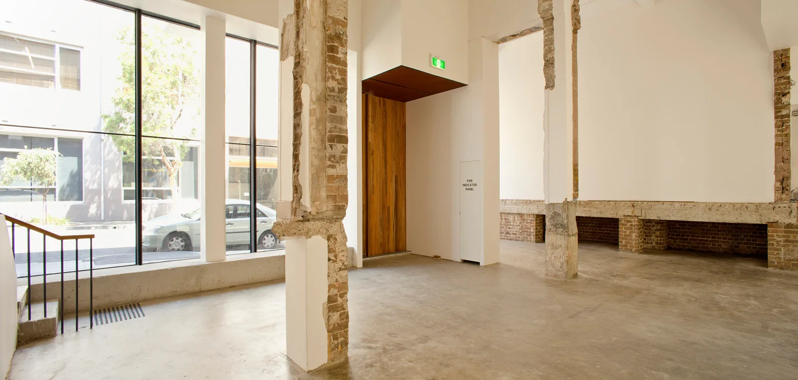 An empty industrial space features smooth concrete flooring, large floor-to-ceiling windows, and raw columns showing exposed brick and concrete.