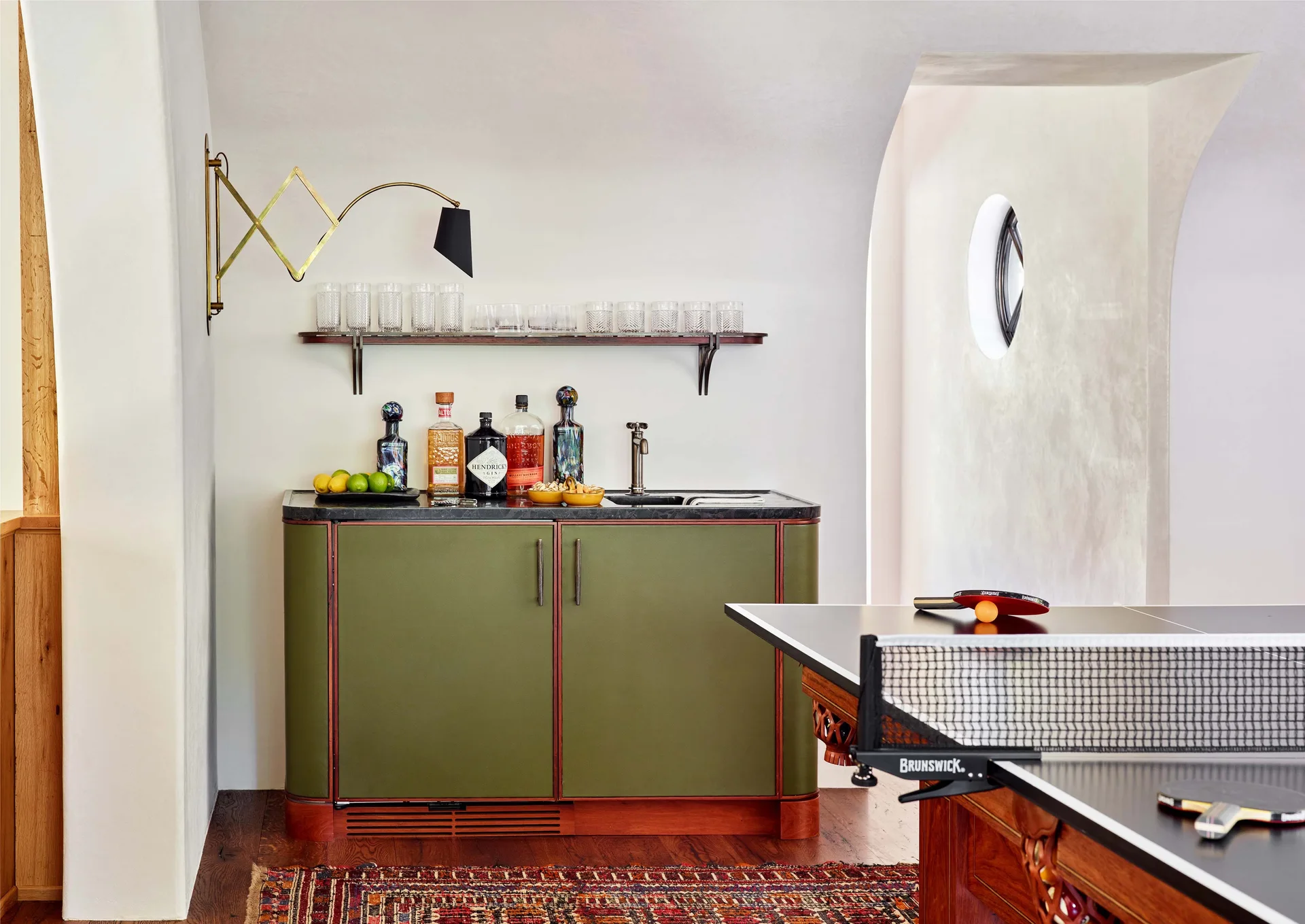 An olive green wet bar with a dark countertop and a brass accordion sconce stands in a game room near a ping pong table.