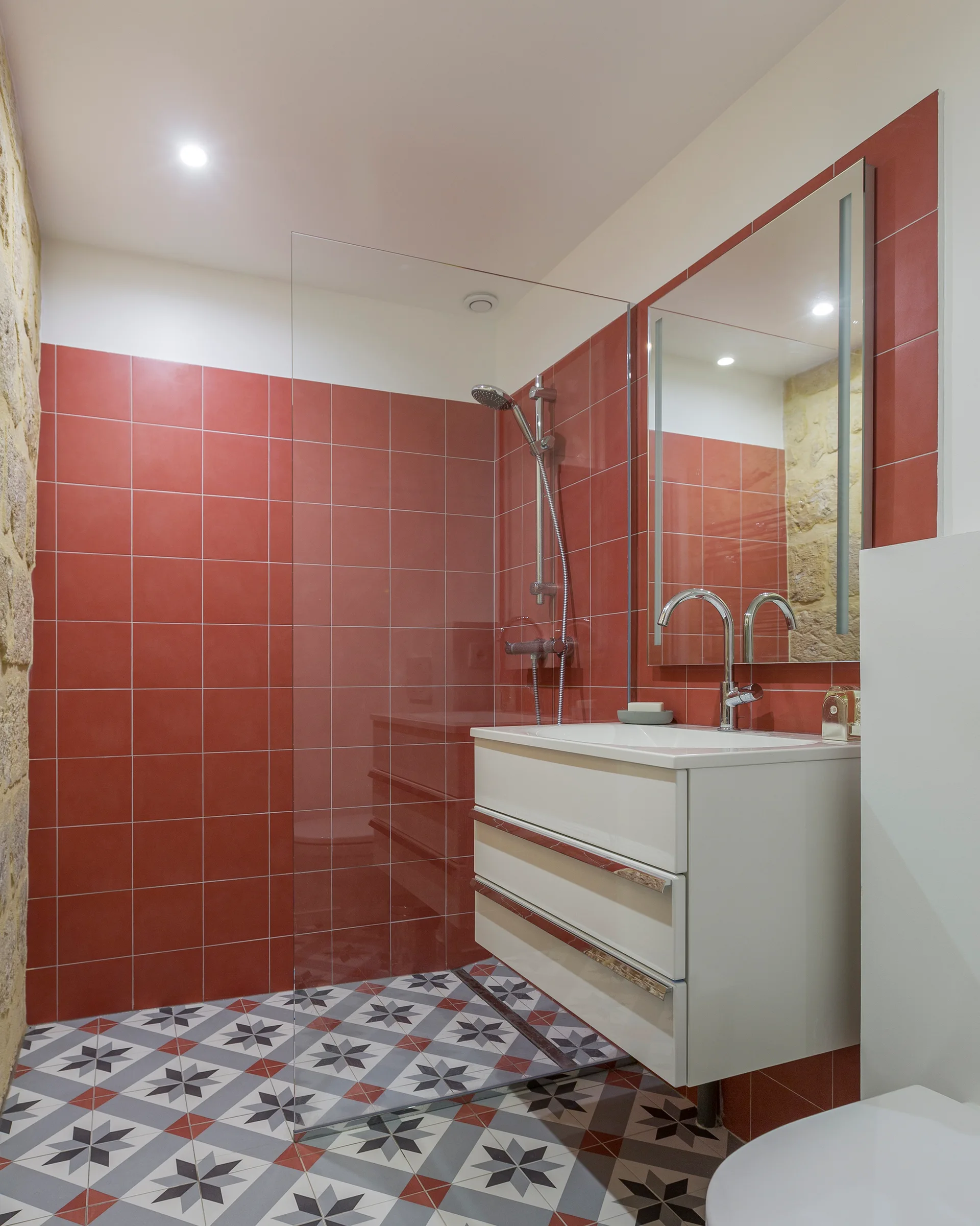 A modern bathroom features glossy red wall tiles, a white vanity with a chrome faucet, and a grey and white geometric star patterned tile floor.