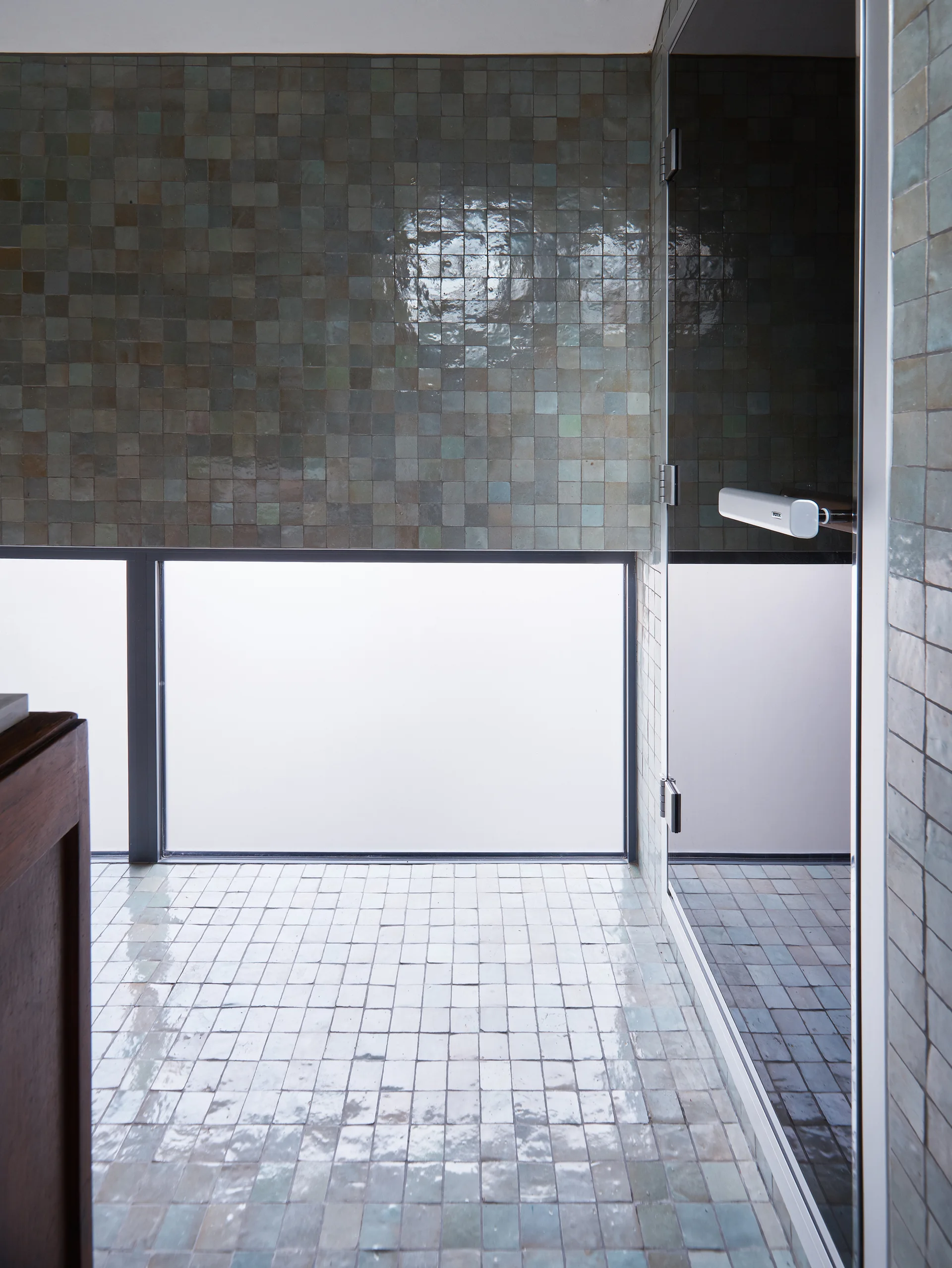 A modern bathroom features glossy green tiled walls and floors, a low rectangular window, and a glass shower door.