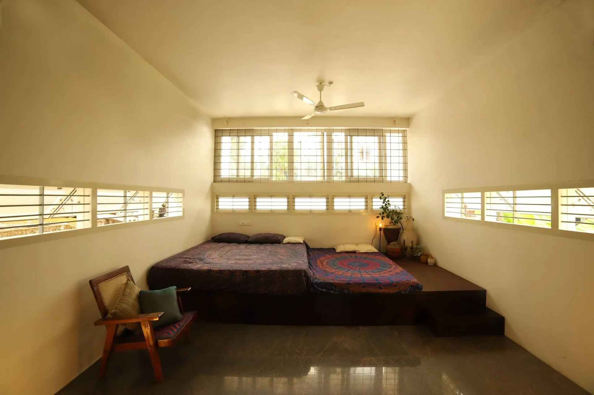 A low-profile wooden platform bed with blue and purple patterned bedding sits beneath horizontal windows in an off-white minimalist bedroom.