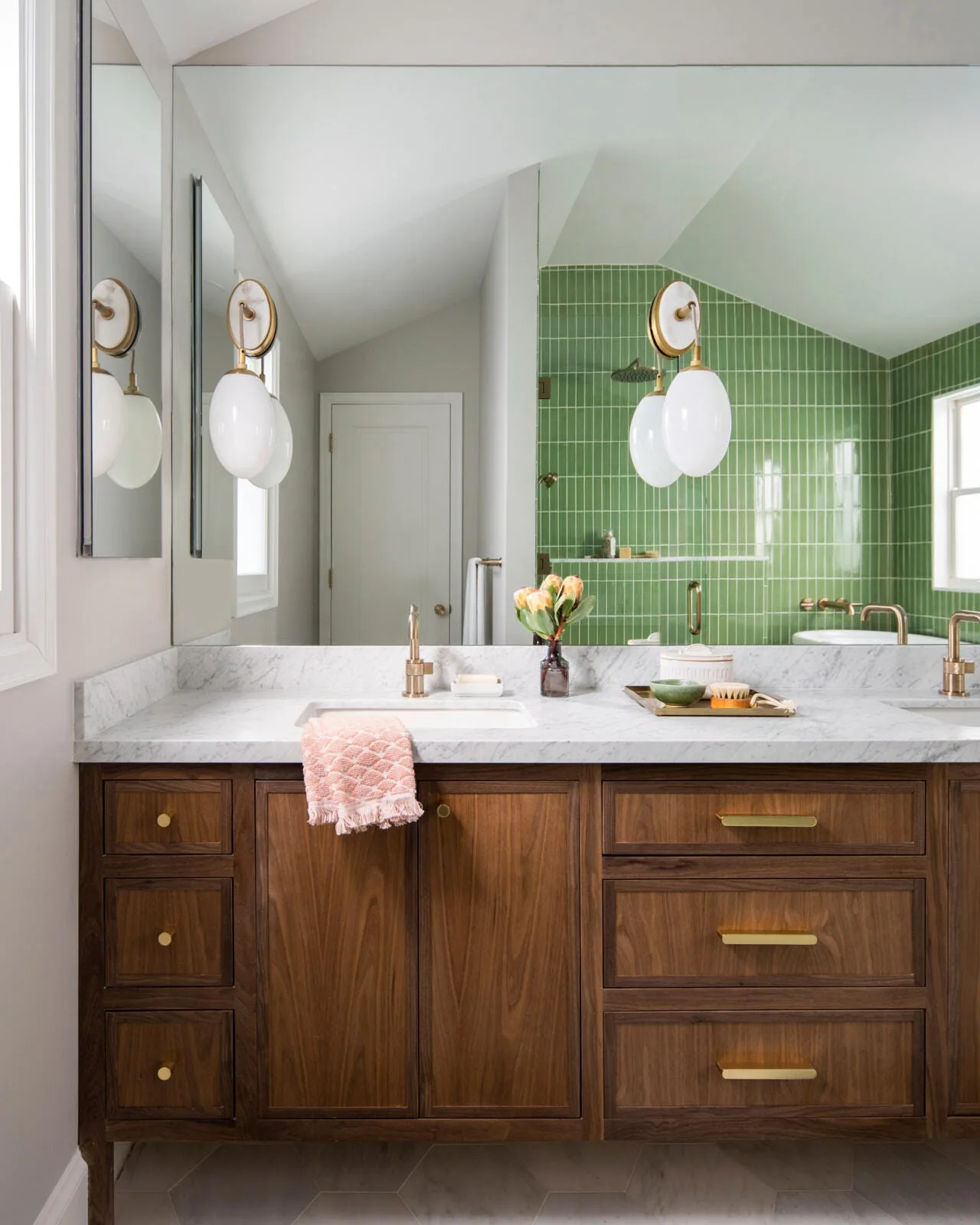 A bathroom features a natural wood vanity with a white marble countertop, brass hardware, and a mirror reflecting a green tiled shower.