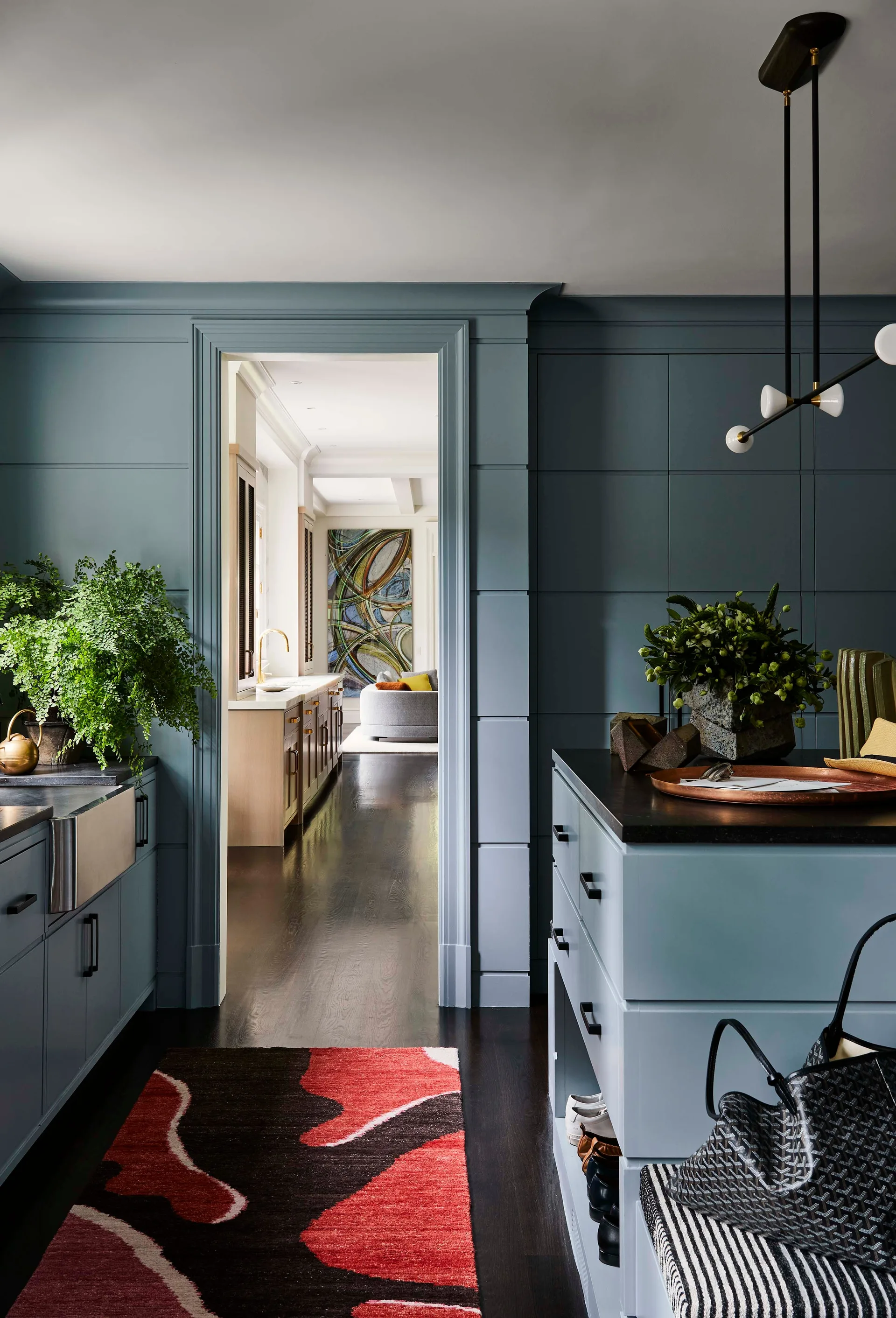 Blue paneled kitchen entryway featuring matching cabinetry, a dark wood floor, and a colorful abstract rug leading toward a sunlit adjoining room.