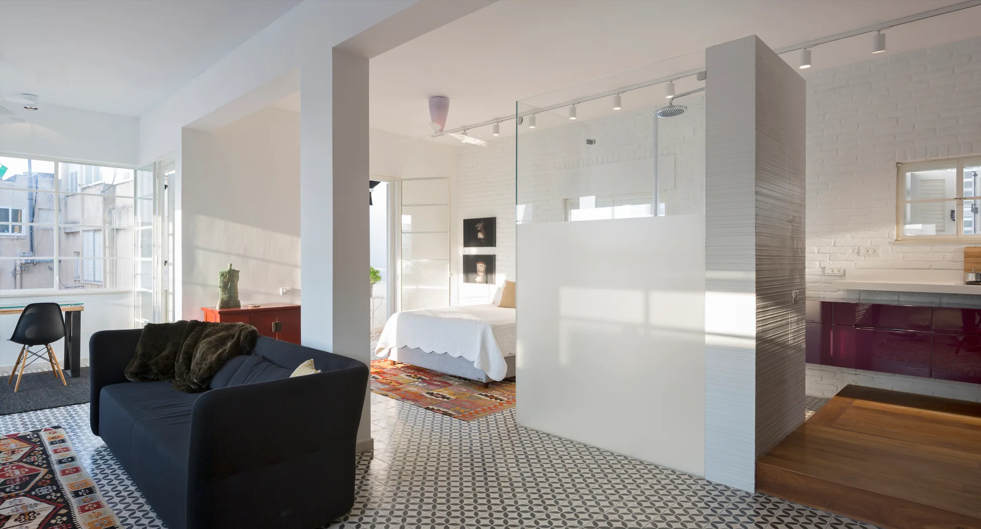 An open-concept studio apartment featuring geometric patterned floor tiles, a dark sofa, and a glass partition separating the bedroom area.