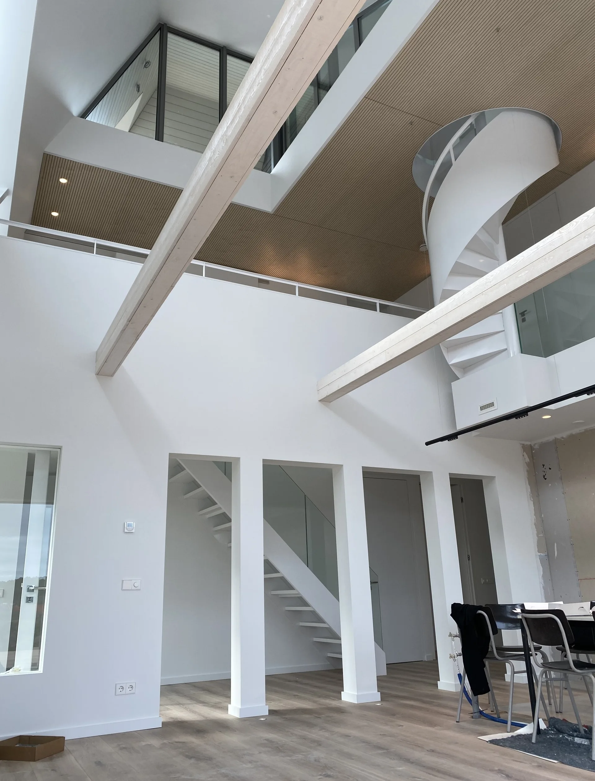 A bright modern loft interior featuring a white spiral staircase, exposed light-wood ceiling beams, a slatted wood ceiling, and white walls.