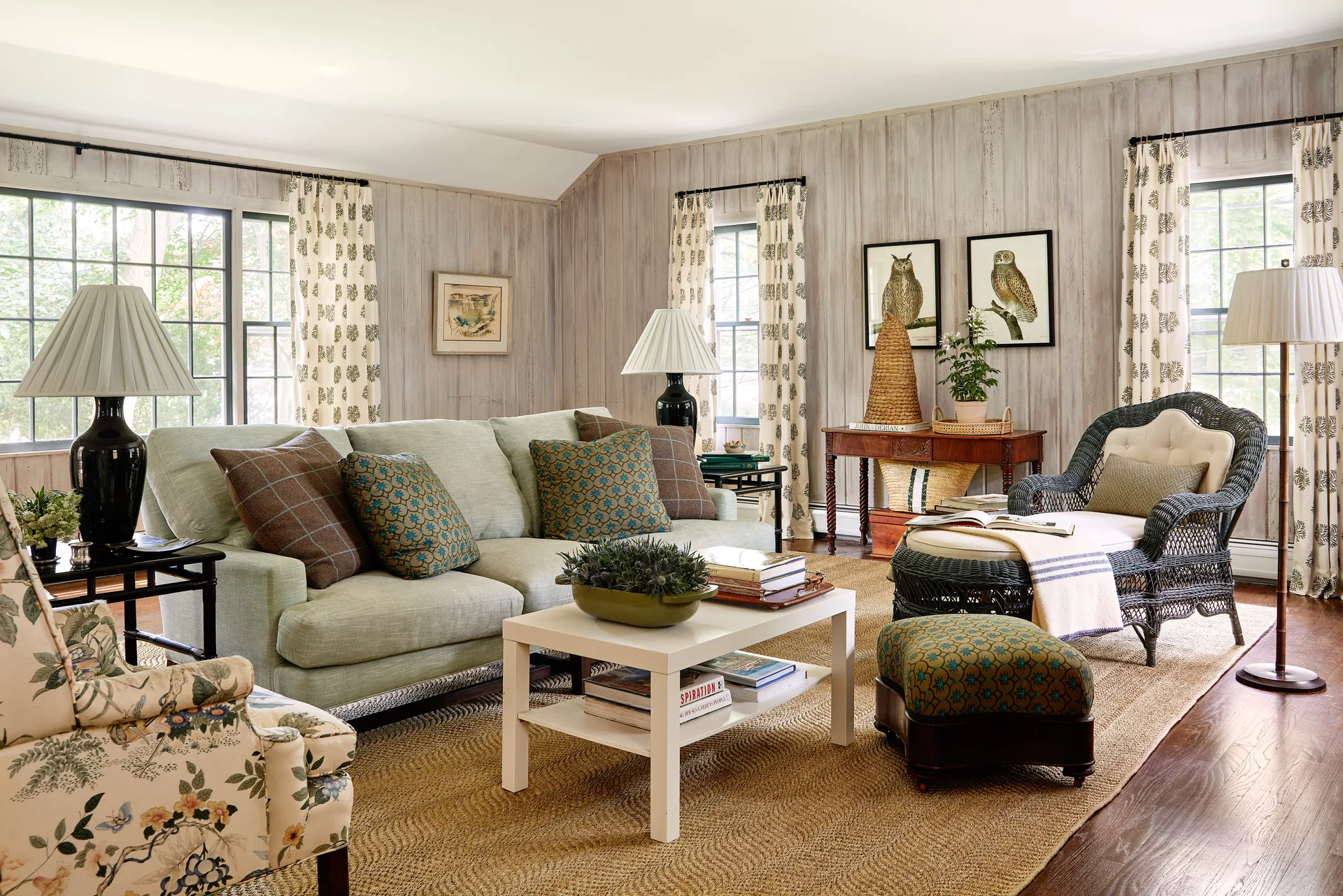 A cottage living room features a light green sofa, whitewashed wood paneled walls, a black wicker lounge chair, and a woven rug.