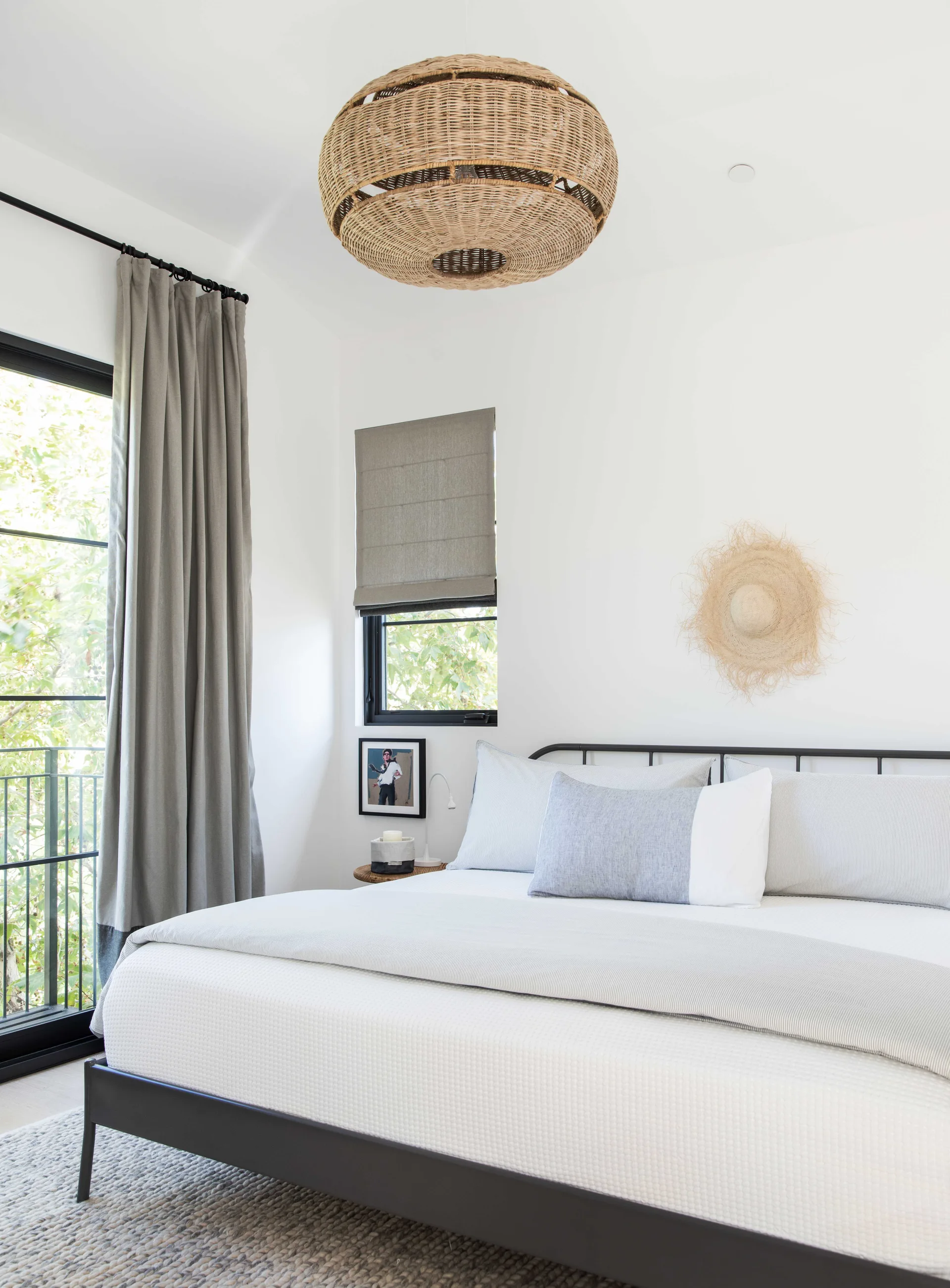 A light-filled bedroom features a black metal bed frame with grey linens beneath a large wicker pendant light.