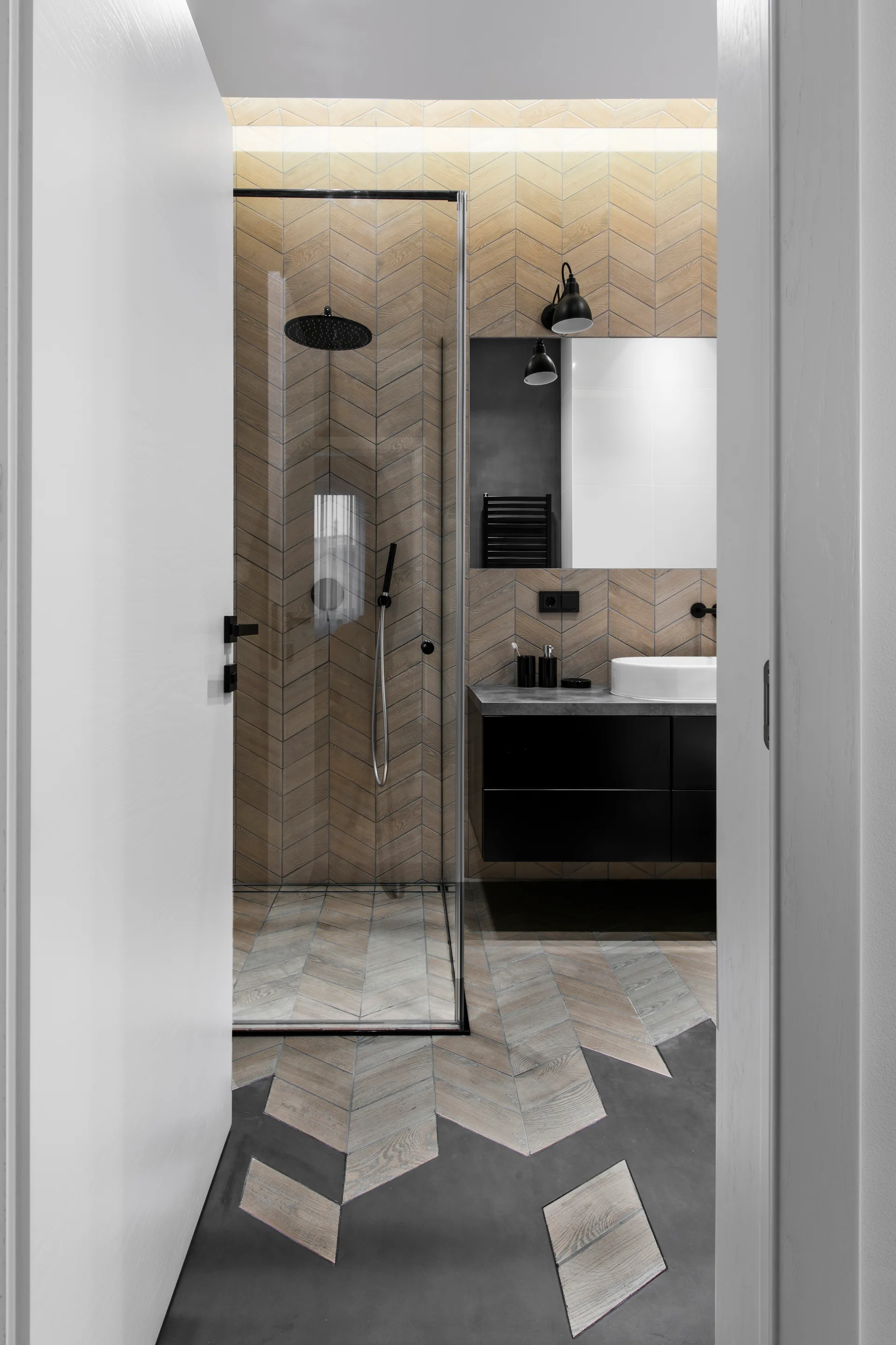 A modern bathroom with chevron wood-look wall tiles, a glass walk-in shower, and a black floating vanity seen through a white doorway.