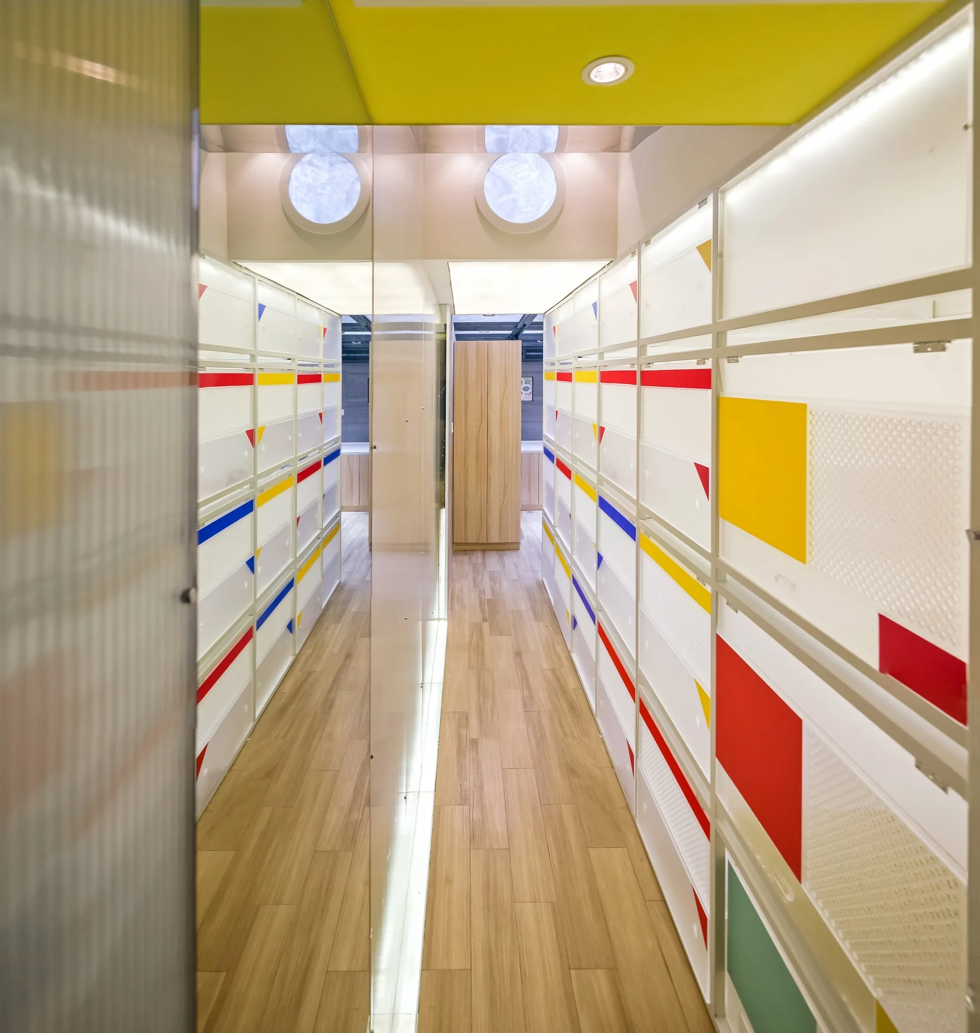A narrow hallway features white modular storage with yellow and red accents, light wood floors, and a floor-to-ceiling mirrored partition.
