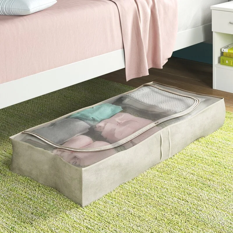 A low-profile beige fabric storage bag with a transparent zippered top sits on a green carpet under a white bed frame.