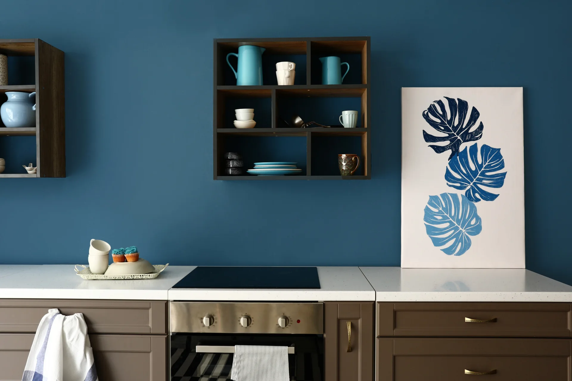 A kitchen features a deep blue wall, brown base cabinets, a white countertop, and wall-mounted dark wood open shelves displaying ceramic dishes.