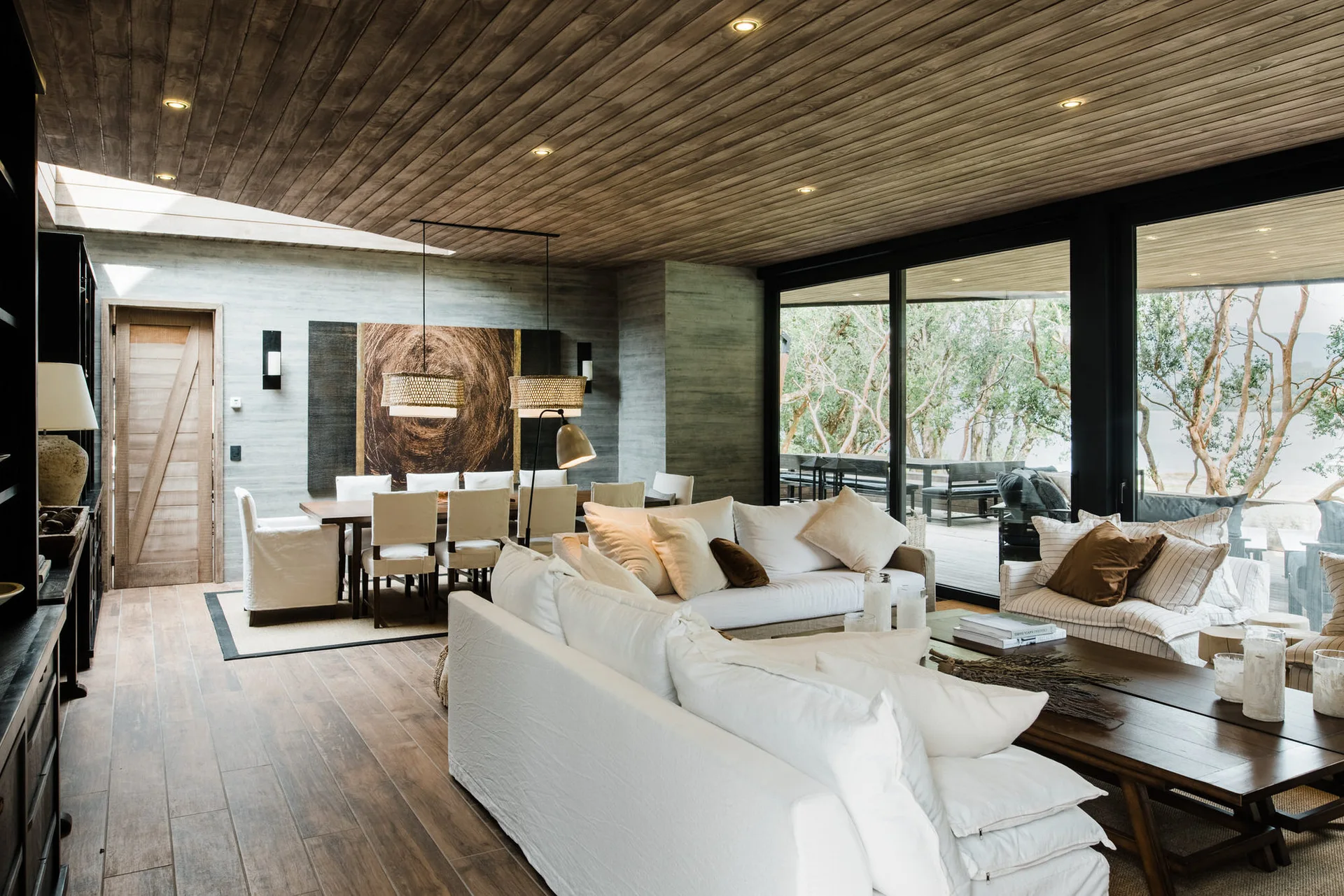 An open-plan living room featuring white sofas, a wooden plank ceiling, a dining table, and large sliding glass doors leading outside.