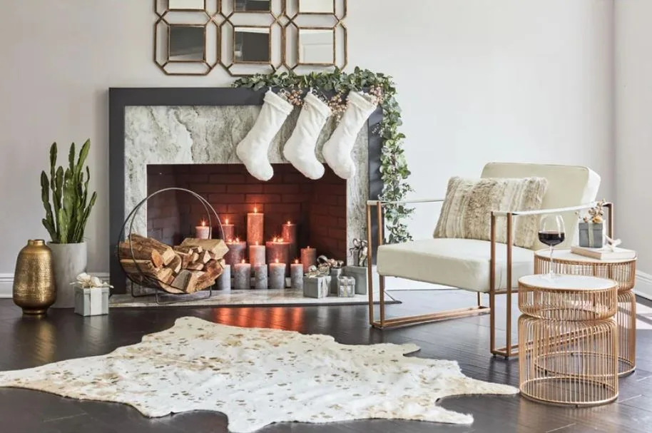 A decorated stone fireplace with white stockings and lit candles sits next to a modern armchair with a gold frame and gold wire side tables.