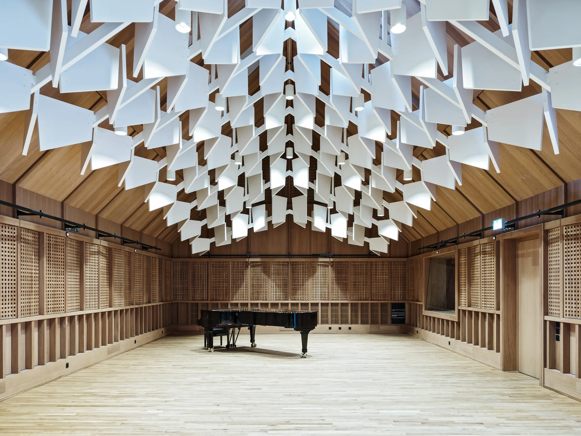 A music performance hall features a black grand piano centered on a wood floor beneath a vaulted timber ceiling hung with white geometric acoustic baffles.