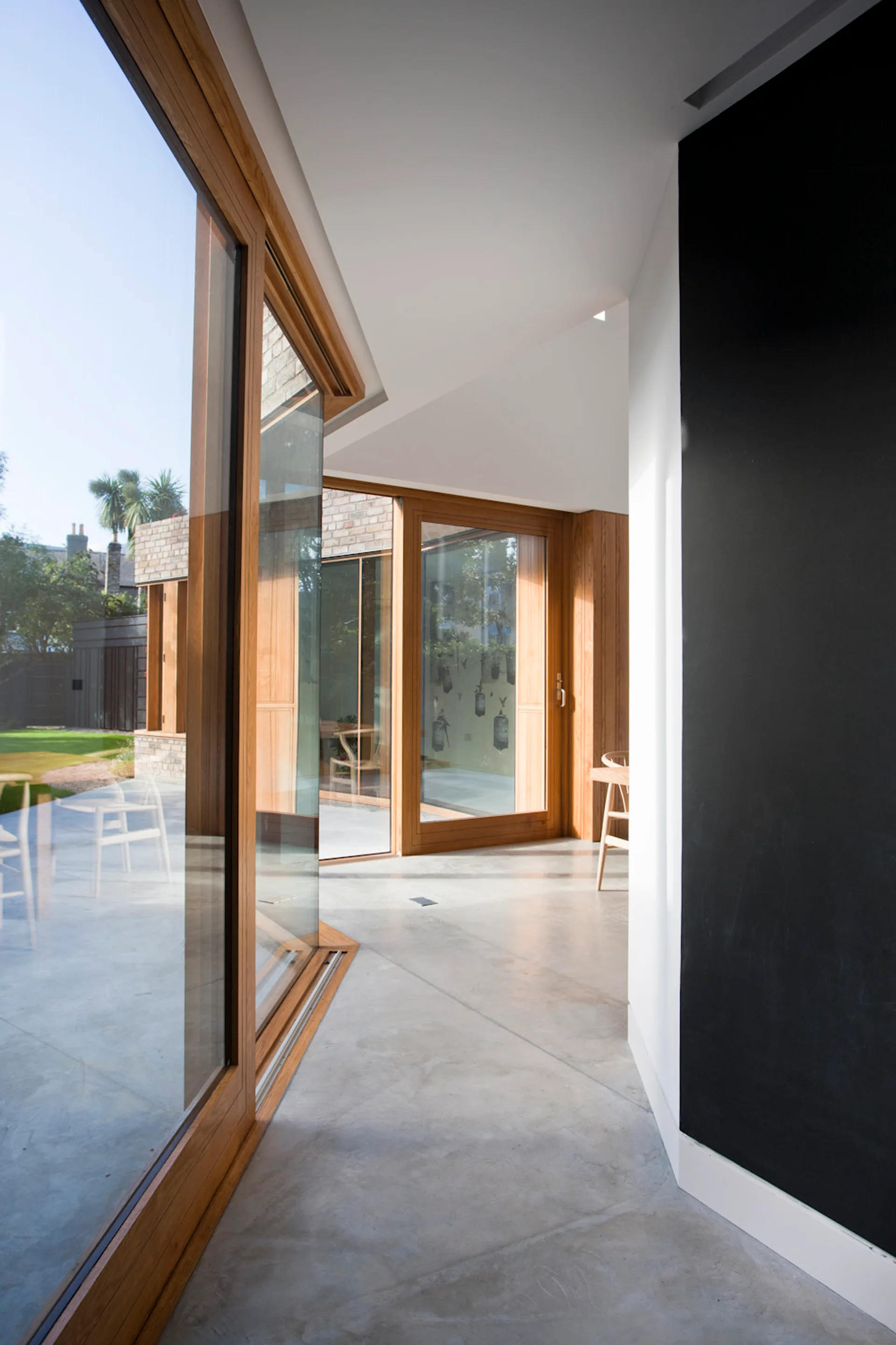A modern hallway features polished concrete flooring, white walls with a black accent section, and large wood-framed glass doors facing a courtyard.