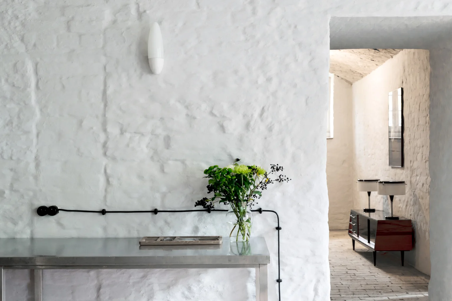 A stainless steel console stands against a heavily textured white plaster wall with exposed black wiring, with a doorway leading to a back hallway.